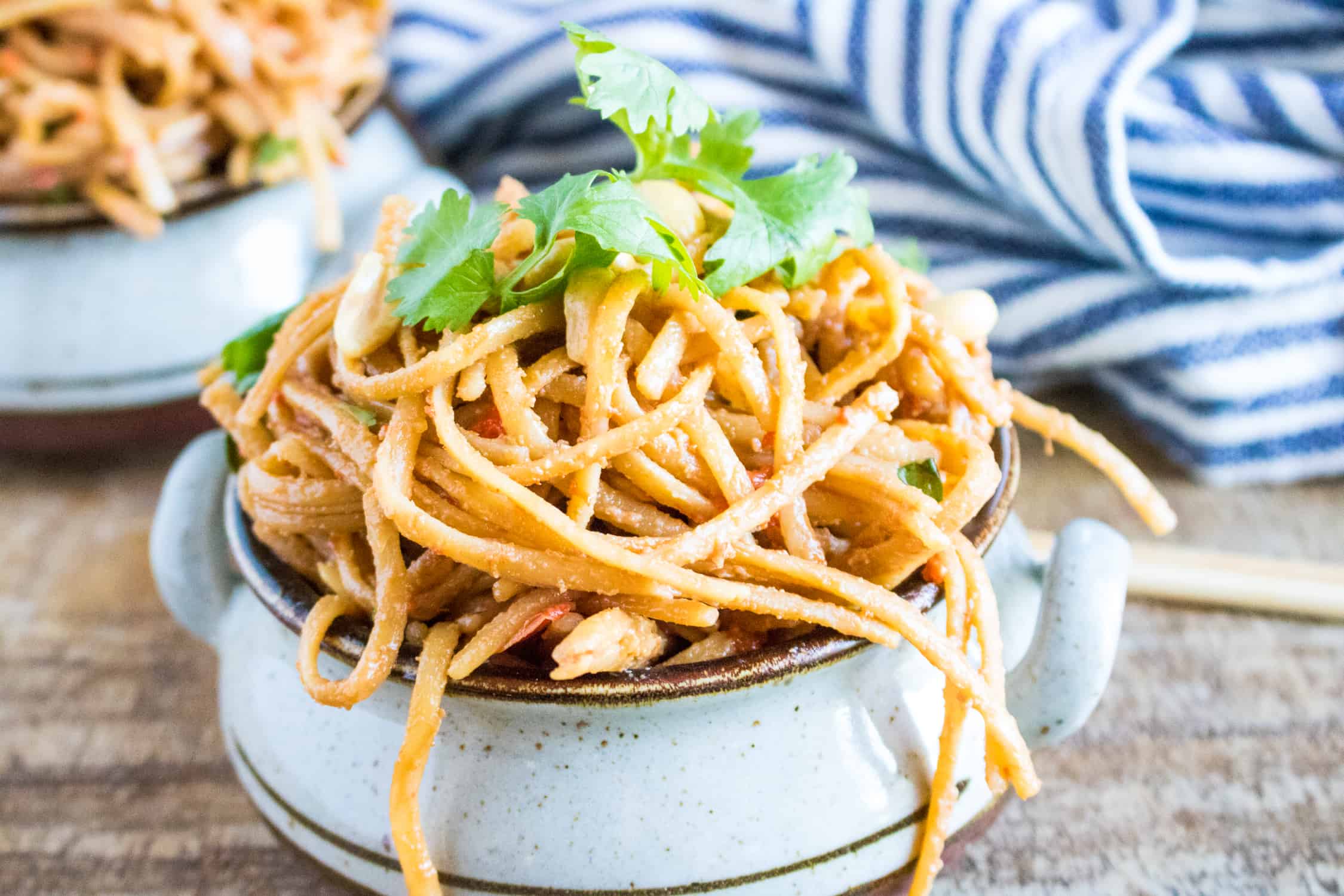 A brown and white ceramic bowl filled with Instant Pot Thai Peanut Noodles garnished with cilantro with a blue and white striped kitchen towel in the background