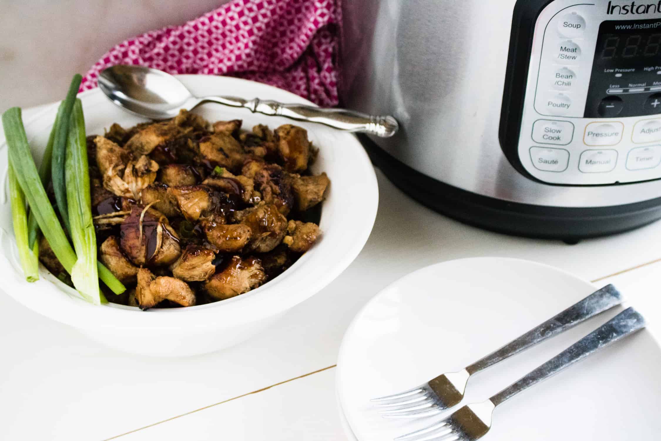 Completed Instant Pot Jerk Chicken Recipe in a white serving bowl placed next to the Instant Pot, two white plates, silver forks, silver serving spoon, and a red print napkin