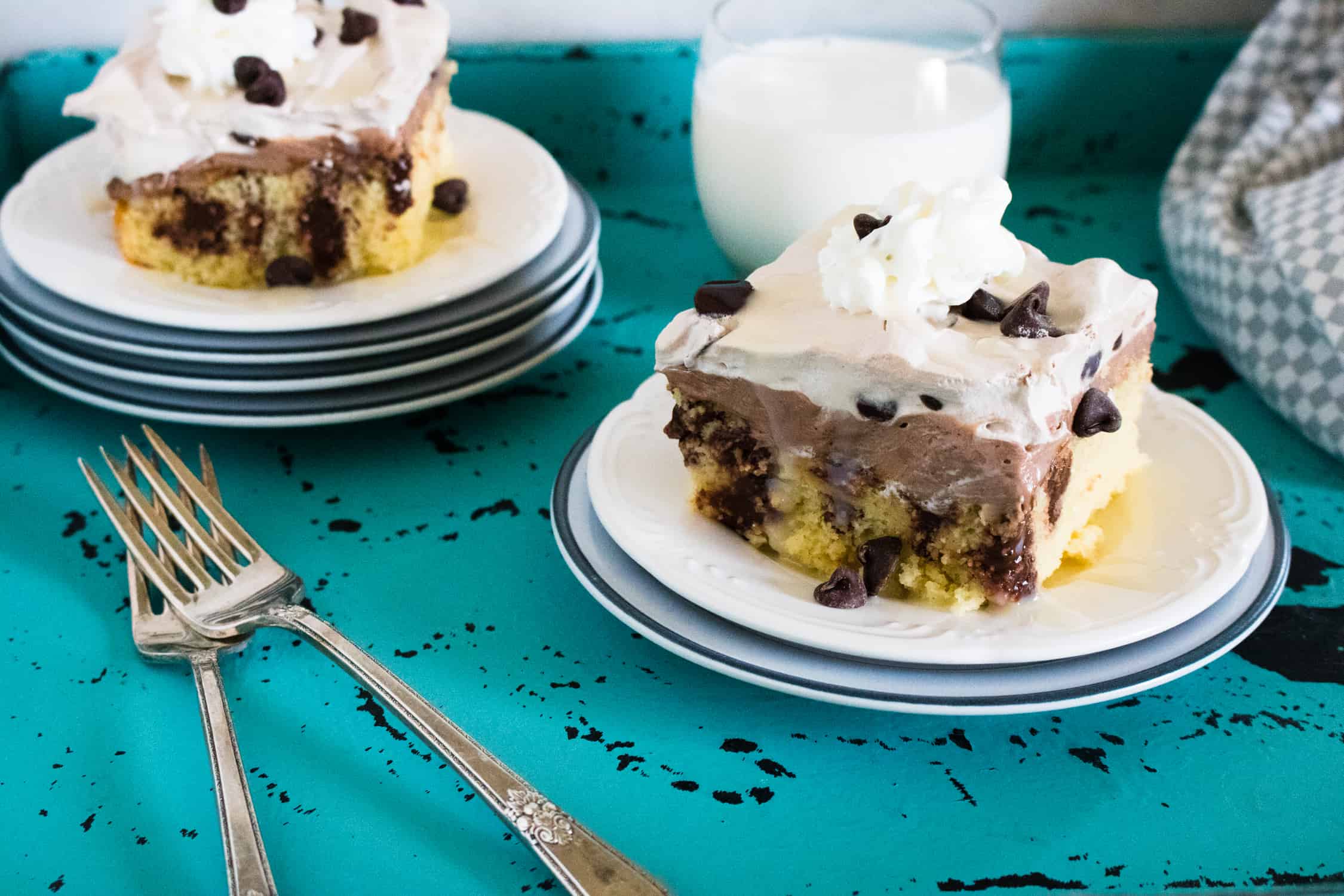 two white plates each with a piece of quadruple chocolate poke cake on them with silver forks in foreground and turquoise tablecloth underneath