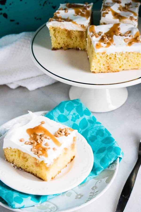 Top-down view of Caramel Toffee Poke Cake on a white cake stand behind a single slice served on a white plate on a blue print napkin