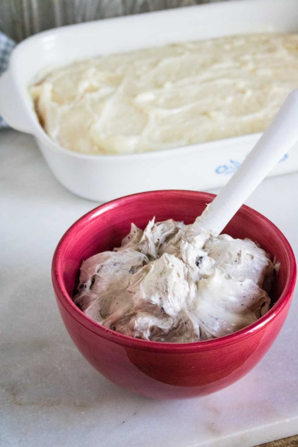Pudding-soaked white cake in a 9x13 baking dish behind a red bowl of Oreo cream cheese filling with a white spoon