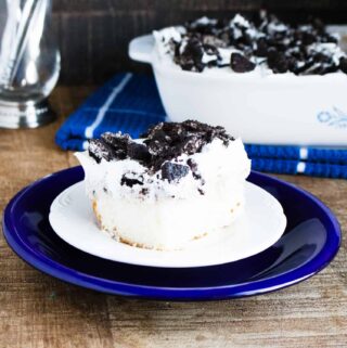 A slice of Oreo Poke Cake served on a white and blue dessert plate and sitting in front of the rest of the Oreo Poke Cake in a white corning ware sheet pan