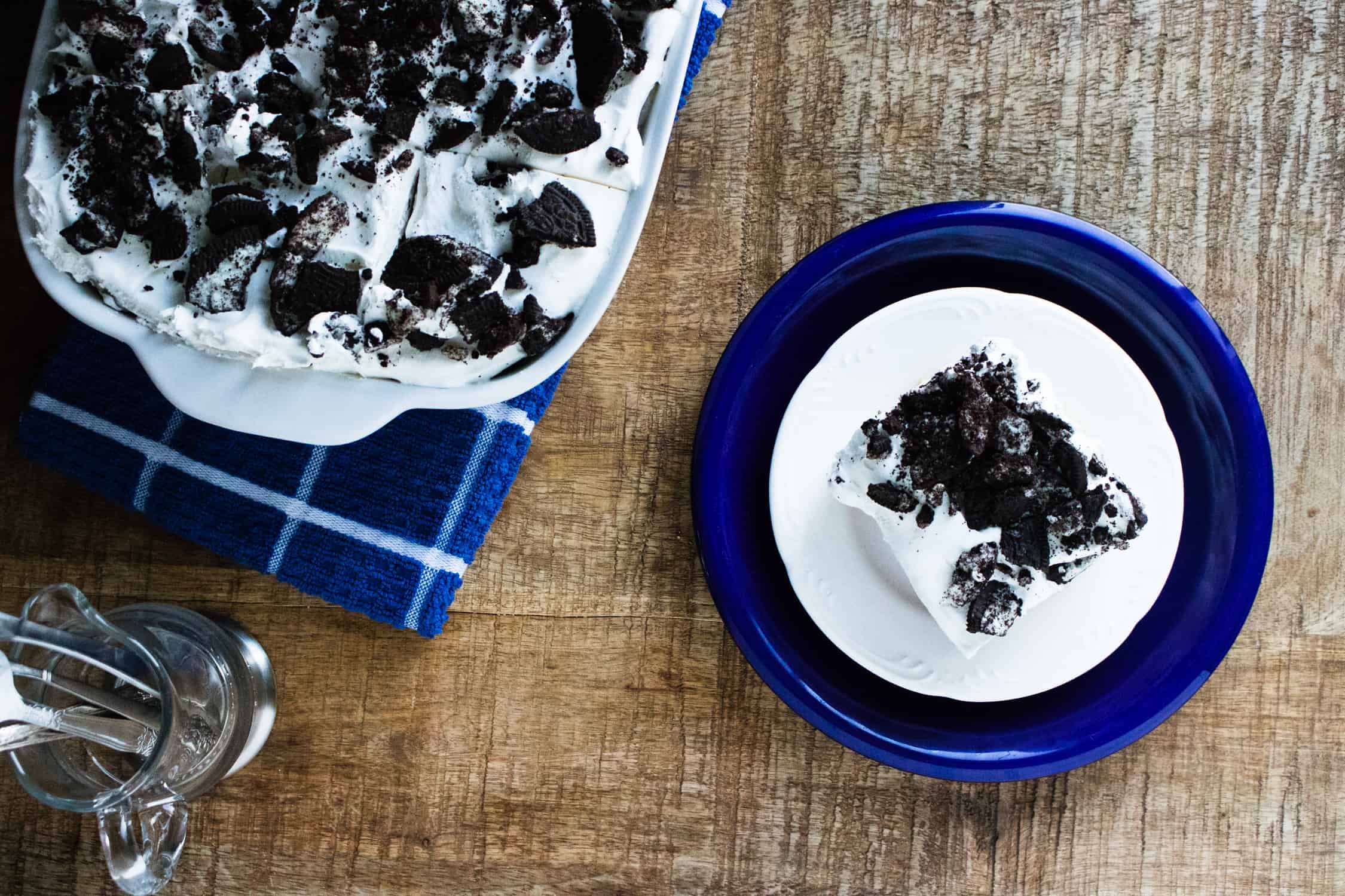 Overhead view of a slice of Oreo poke cake on a blue plate next to the baking dish of cake on a blue checked towel