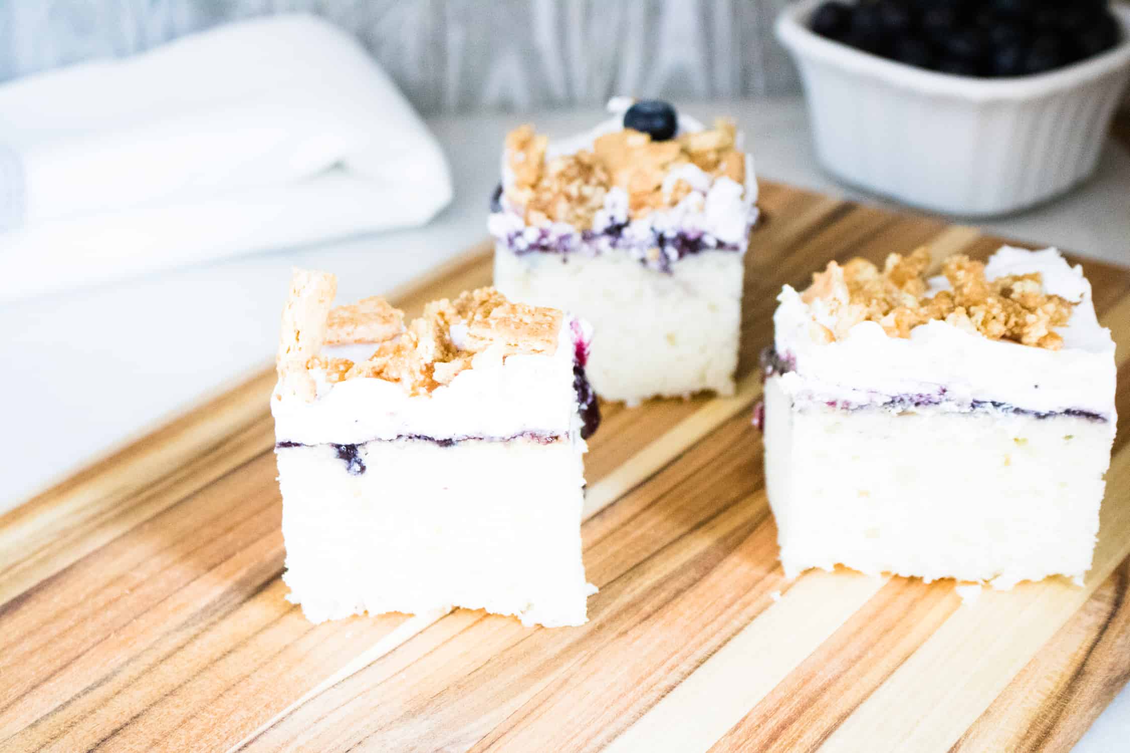 Three slices of blueberry buckle poke cake on a wooden cutting board in front of a white napkin and white bowl of blueberries