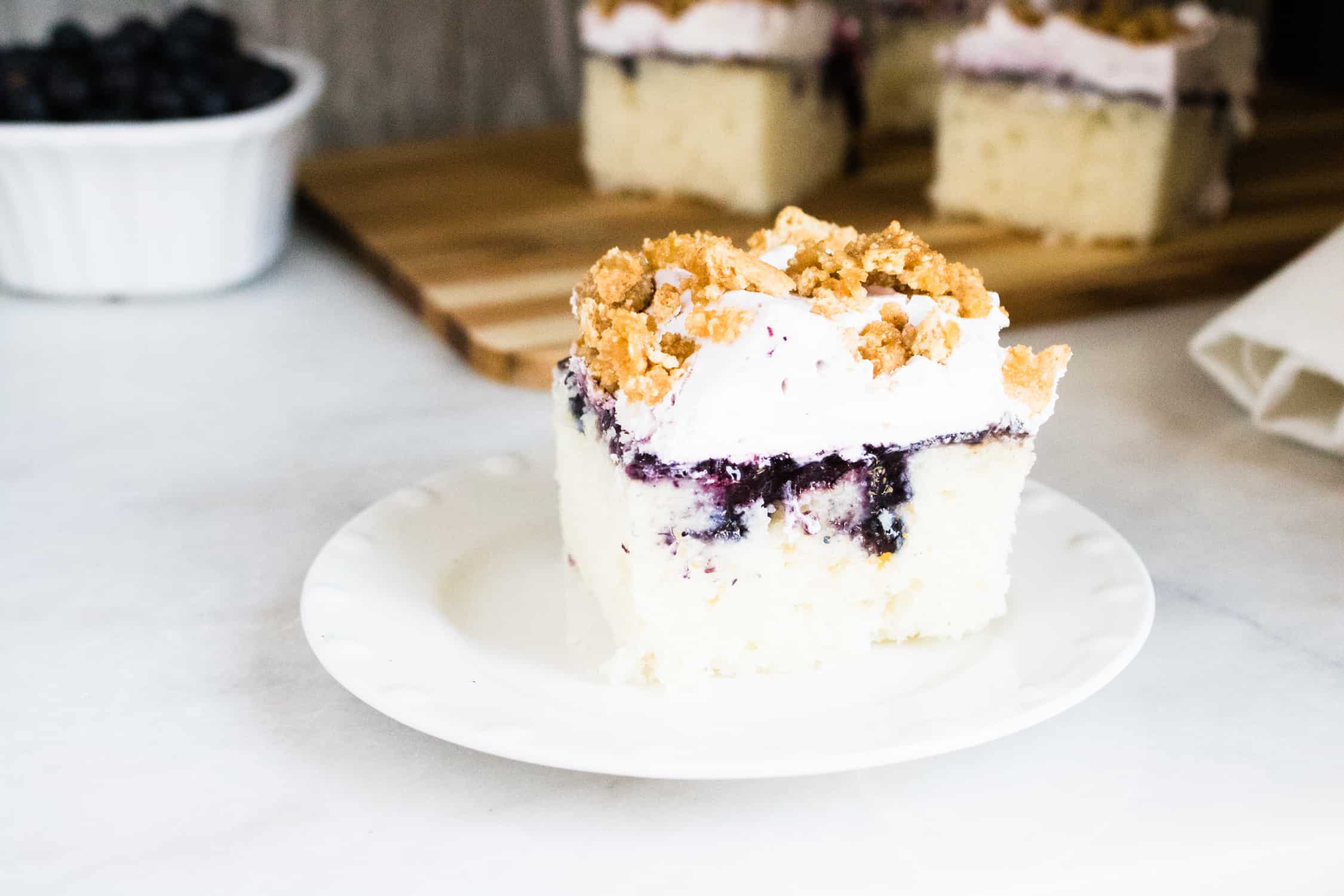 one slice of blueberry buckle poke cake served on a white dessert plate sitting in front of two slices on a wooden cutting board next to a bowl of blueberries