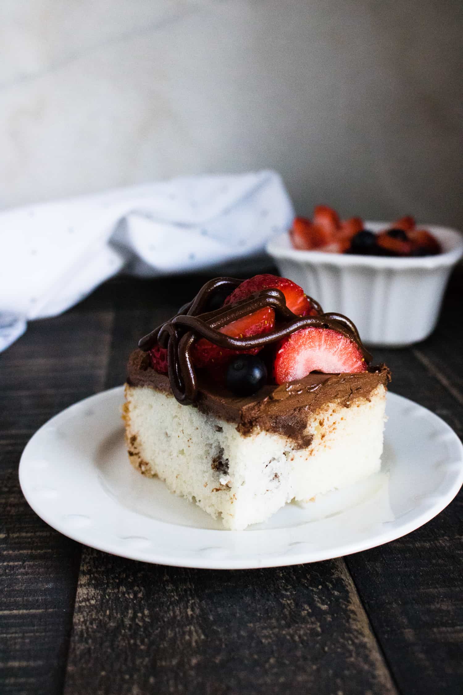 one slice of chocolate ganache poke cake with berries served on a white plate set in front of a white cloth napkin and white bowl of mixed berries