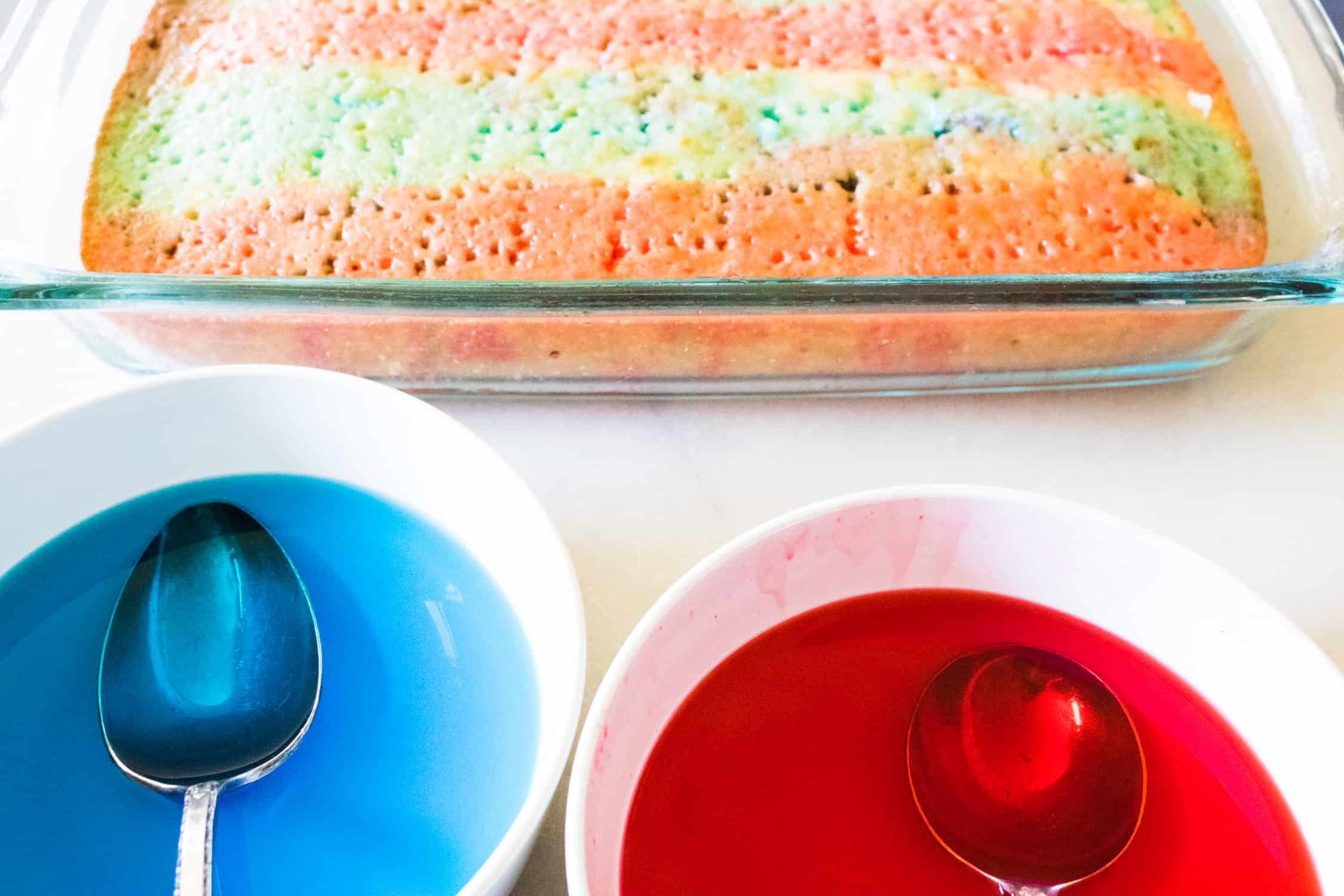 prepared white sheet cake inside a baking dish next to a bowl of blue gelatin and a bowl of red gelatin
