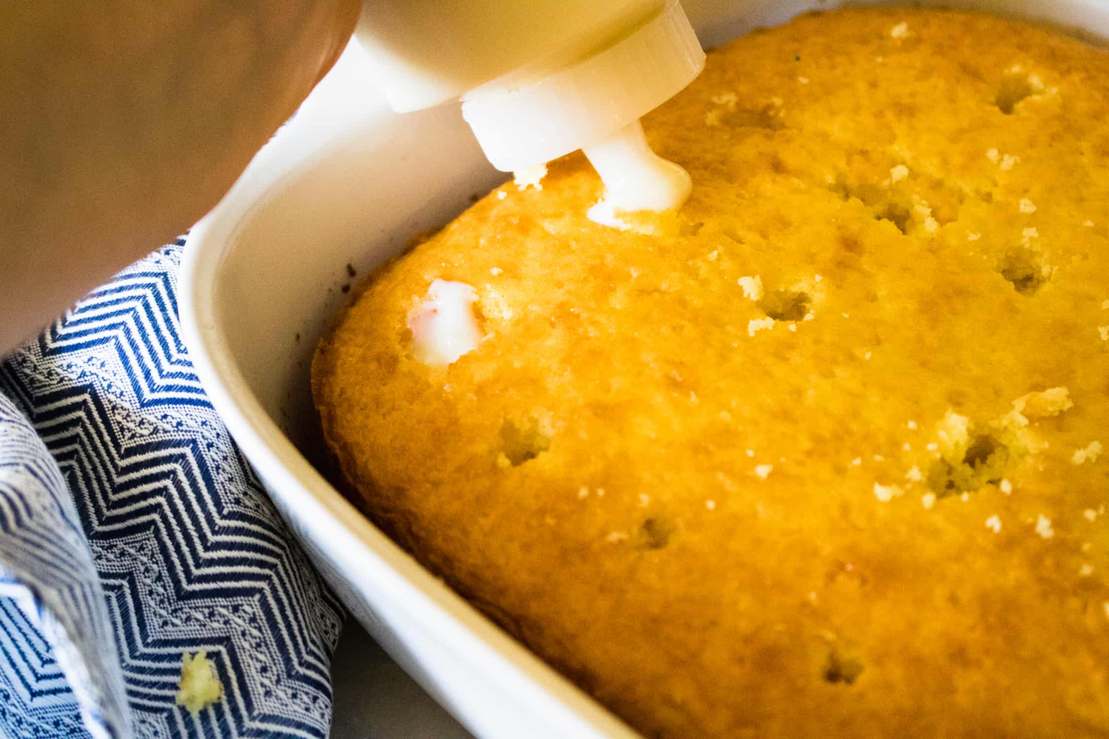 Lemon pudding being poured into the wooden-spoon holes of a lime poke cake in a white baking dish