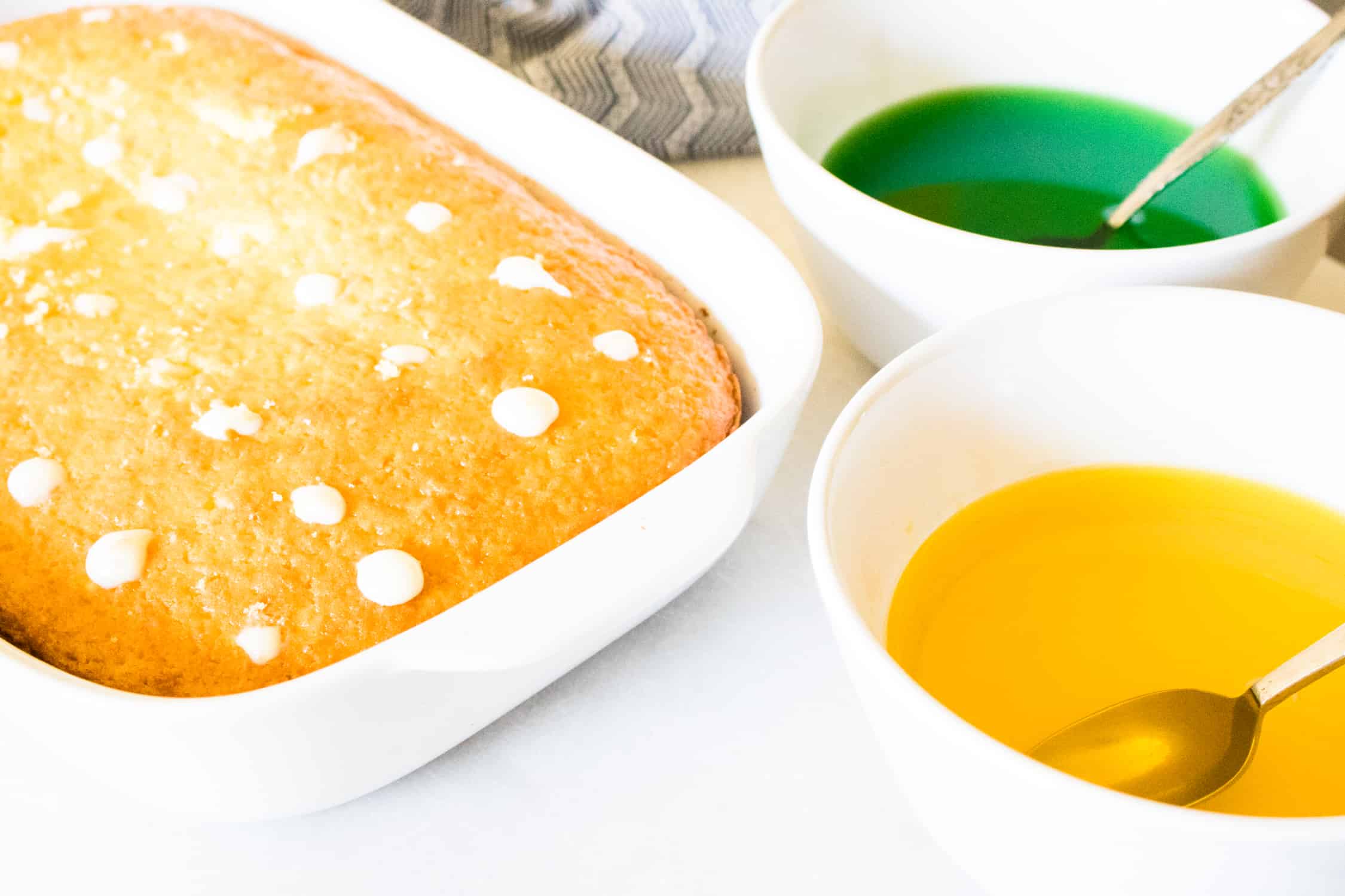 Lemon Lime Poke Cake in a white cake pan next to a white bowl of lime gelatin and a white bowl of lemon gelatin