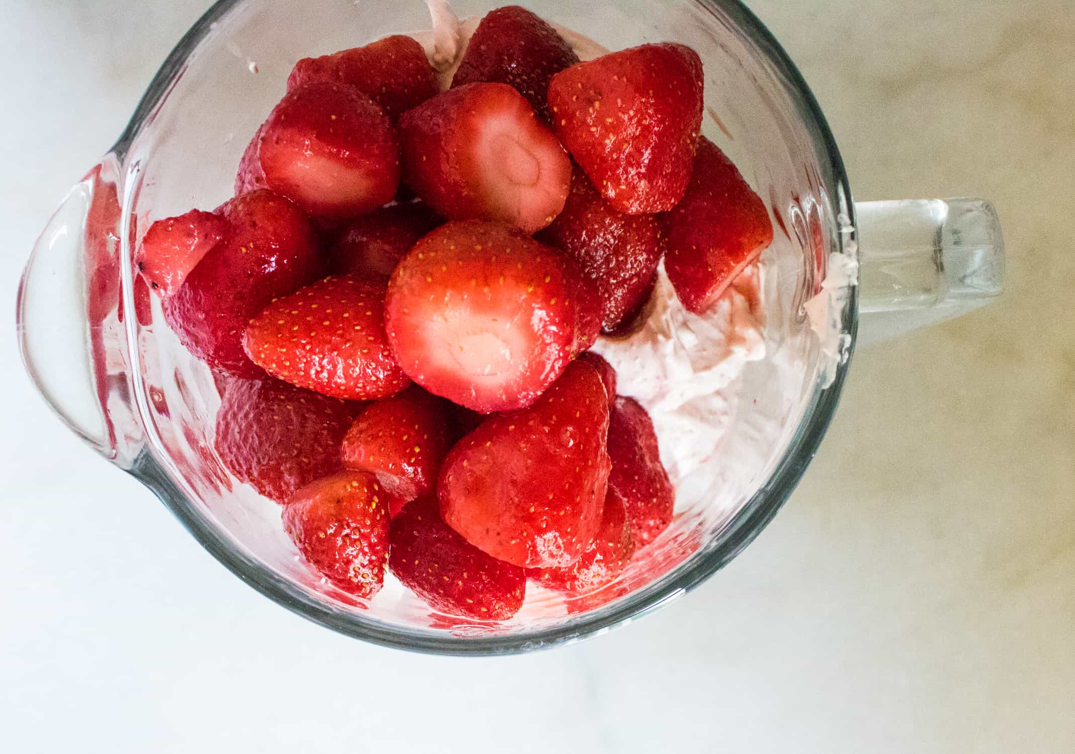top-down view of a clear glass blender pitcher filled with cream cheese, milk, fresh strawberries and red velvet pudding mix