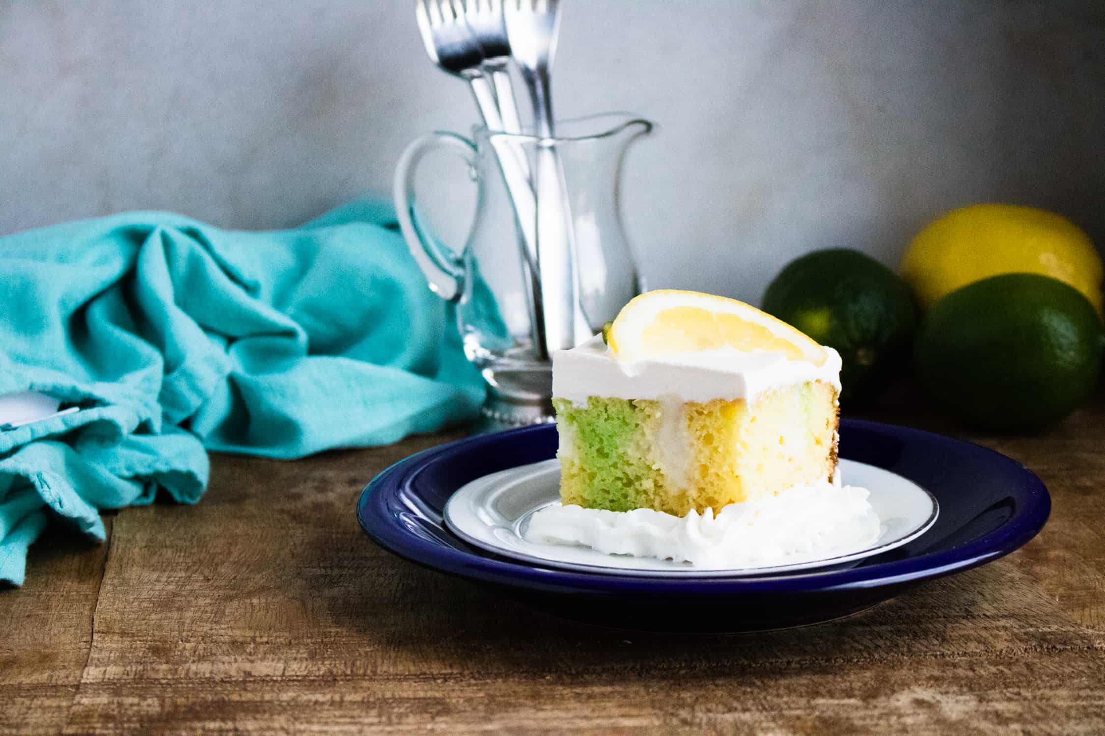 a slice of Lemon Lime Poke Cake on a white saucer on top of a blue plate with a teal kitchen towel, lemons and limes and utensils in a glass pitcher in the background