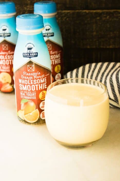 Three bottles of Live Real Farms healthy smoothies next to a glass filled with pineapple orange mango smoothie set in front of a black and white striped kitchen towel 