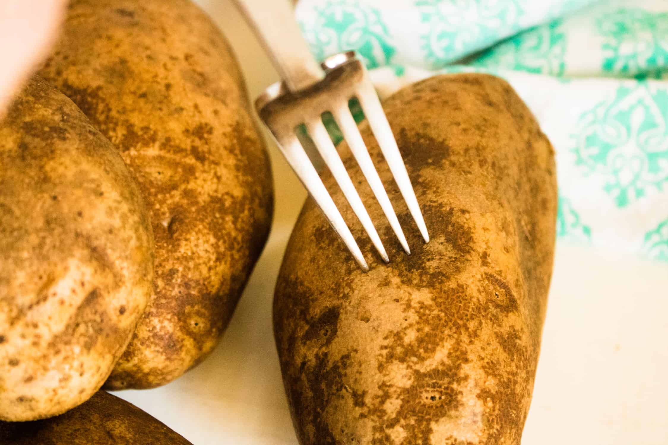 2 potatoes in foreground with one potato being pierced by a silver fork. there is a green and white towel in the background