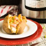 A completed Instant Pot Baked Potato topped with diced ham and melted cheese, served on a white plate with a red charger