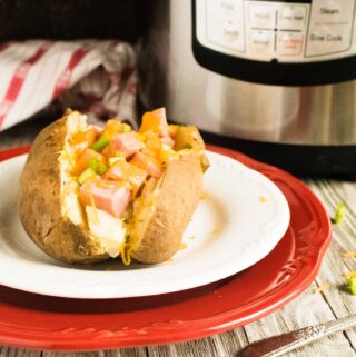 A completed Instant Pot Baked Potato topped with diced ham and melted cheese, served on a white plate with a red charger