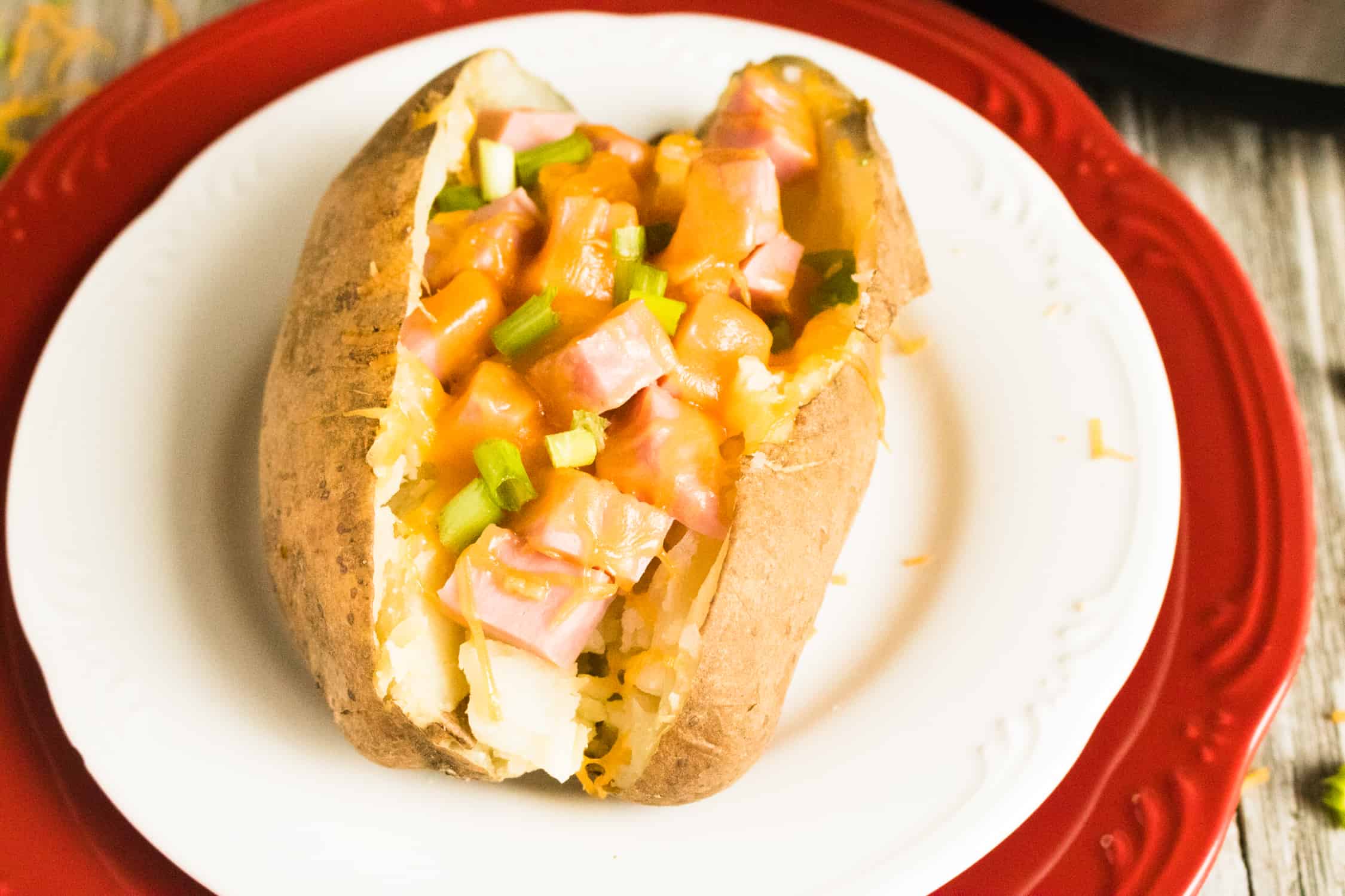 baked potato filled with ham and cheese sitting on a white plate with a red liner plate below,