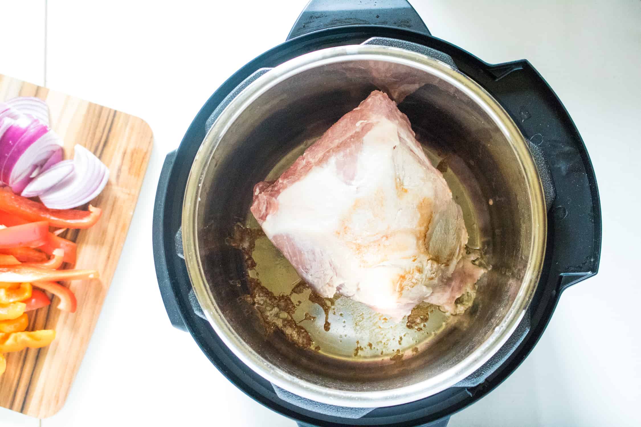 top-down view of pork roast being sauted in the Instant Pot, next to a cutting board with chopped onion and bell peppers on top