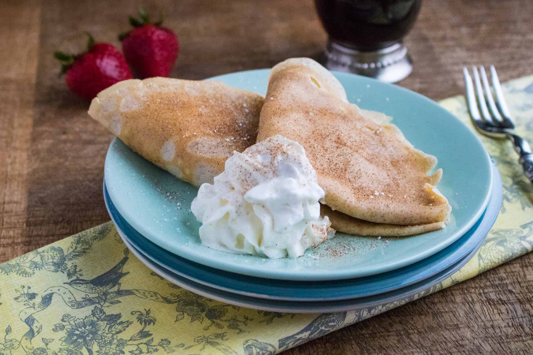 Completed snickerdoodle crepes recipe shows two crepes with whipped cream served on a stack of blue plates sitting on a yellow and blue toile print fabric, along side two strawberries, a clear glass pitcher of maple syrup, and a silver spoon.