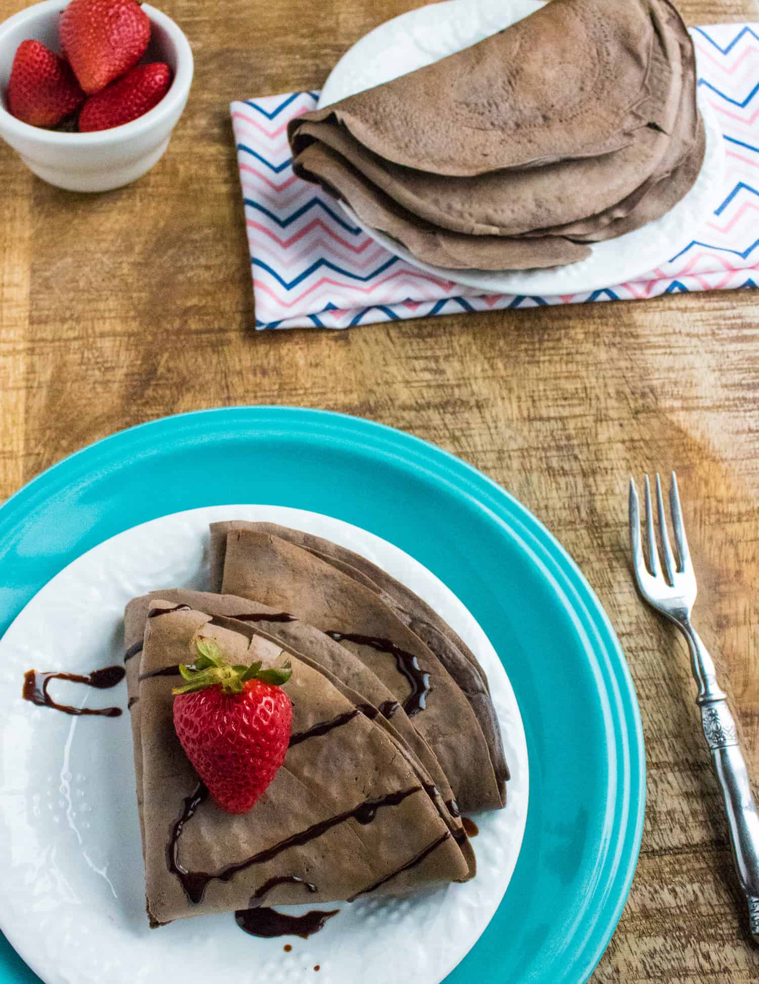 chocolate dessert crepes topped with a strawberry and chocolate sauce, served on a white plate placed on top of a blue plate sitting next to a fork, bowl of strawberries, and a plate of extra crepes