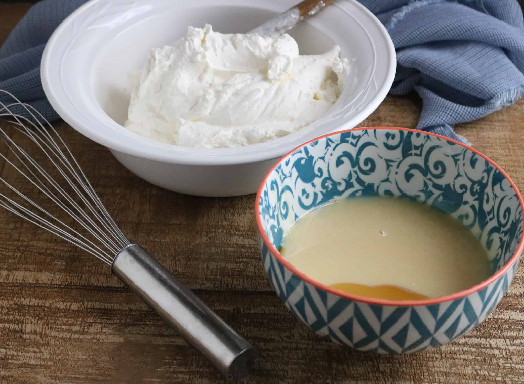 Thick heavy whipping cream in a white bowl and sweetened condensed milk in a teal patterned bowl on a wood table next to a silver whisk and blue kitchen towel