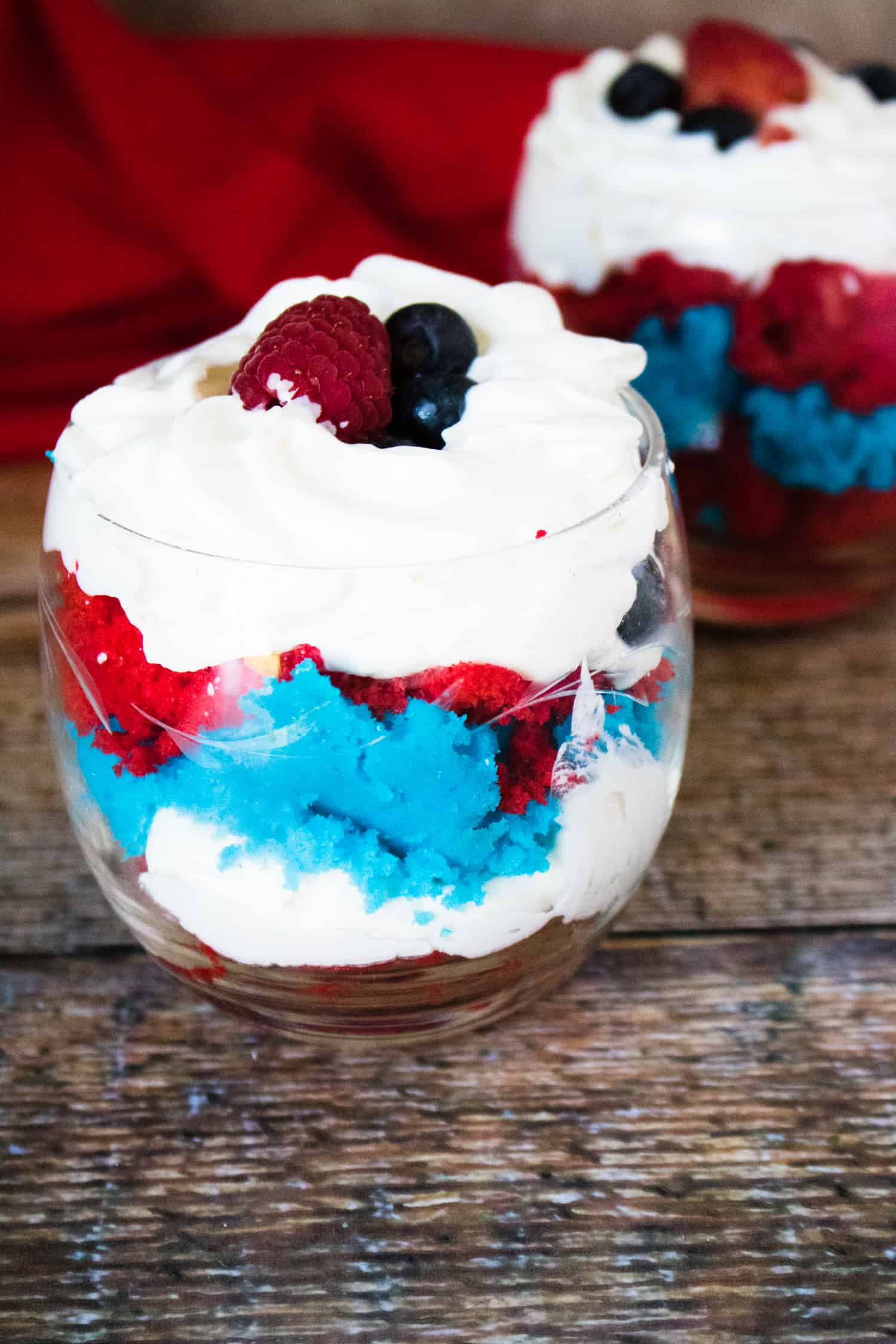 Close-up view of one of these 4th of July Desserts in a cut-glass cup, with a second portion of 4th of July Mini Cake Trifle and a red cloth napkin shown in the background