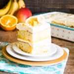 Slice of Tropical Poke Cake serves on a white and orange dessert plate set on top of a blue print napkin with a silver fork