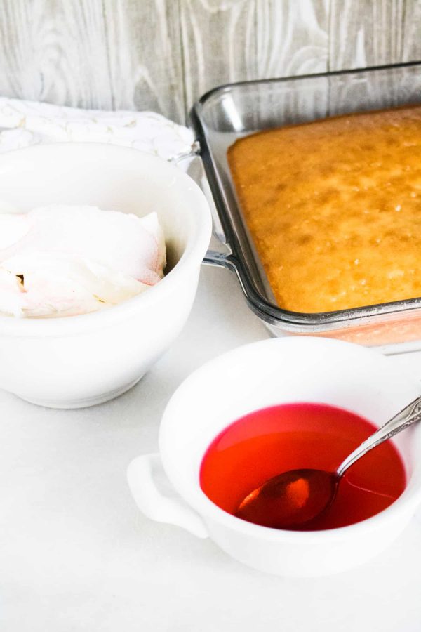 Baked white cake in a glass baking dish next to a bowl of cream cheese and a bowl of red watermelon gelatin liquid
