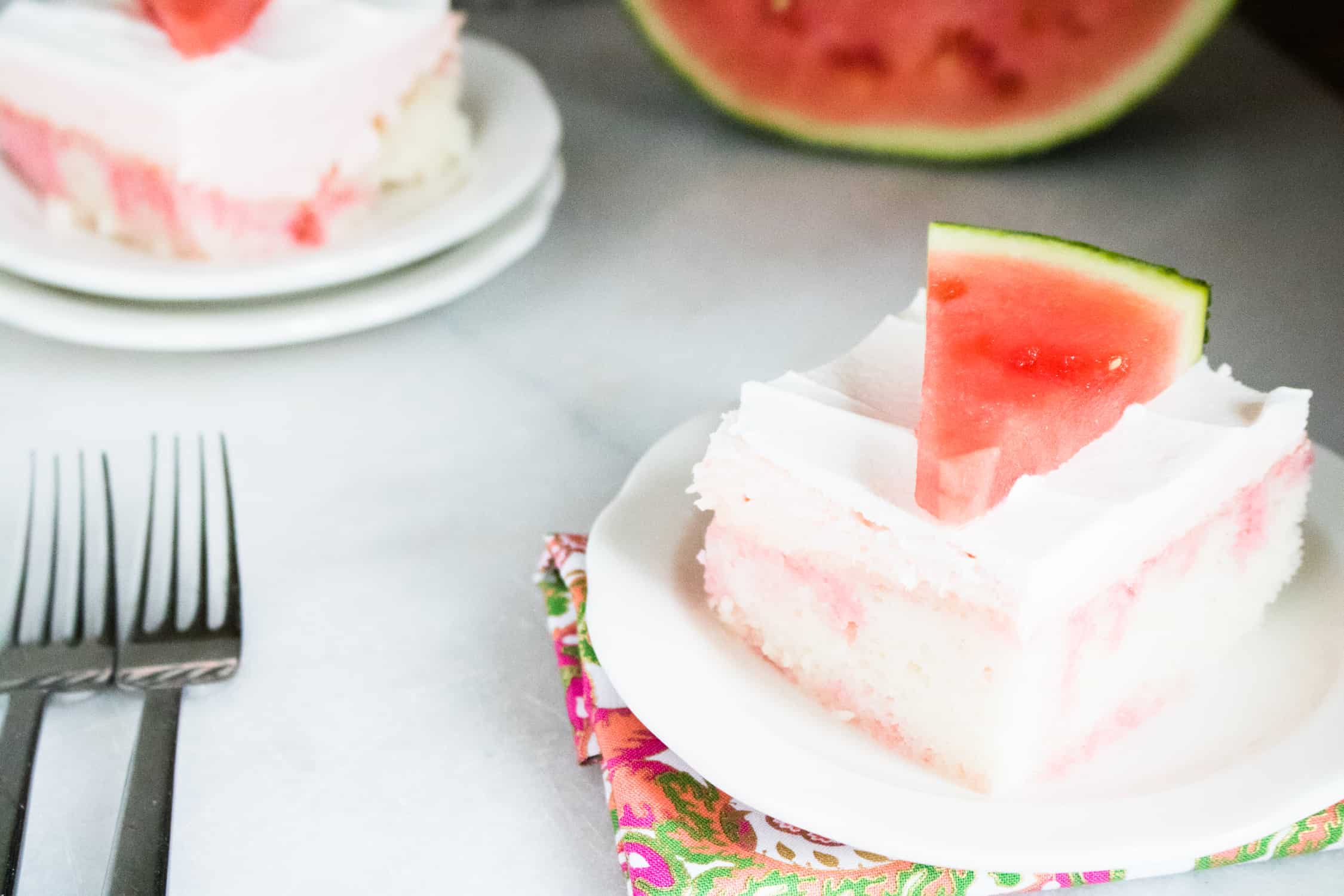 close up view of two servings of Jolly Rancher Watermelon poke cake on white plates and topped with fresh watermelon wedges with two silver forks