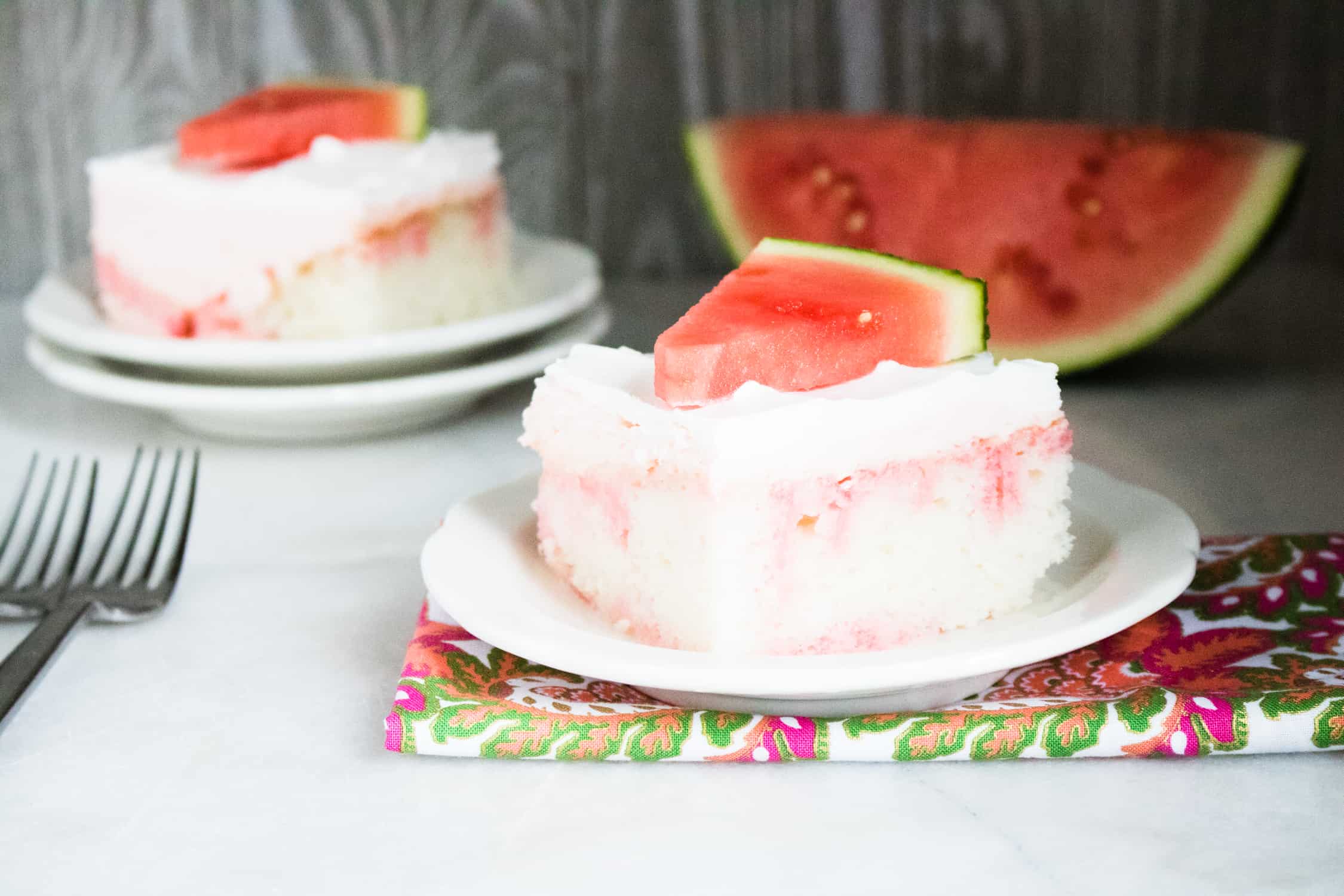 two servings of Jolly Rancher Watermelon poke cake on white plates and topped with fresh watermelon wedges