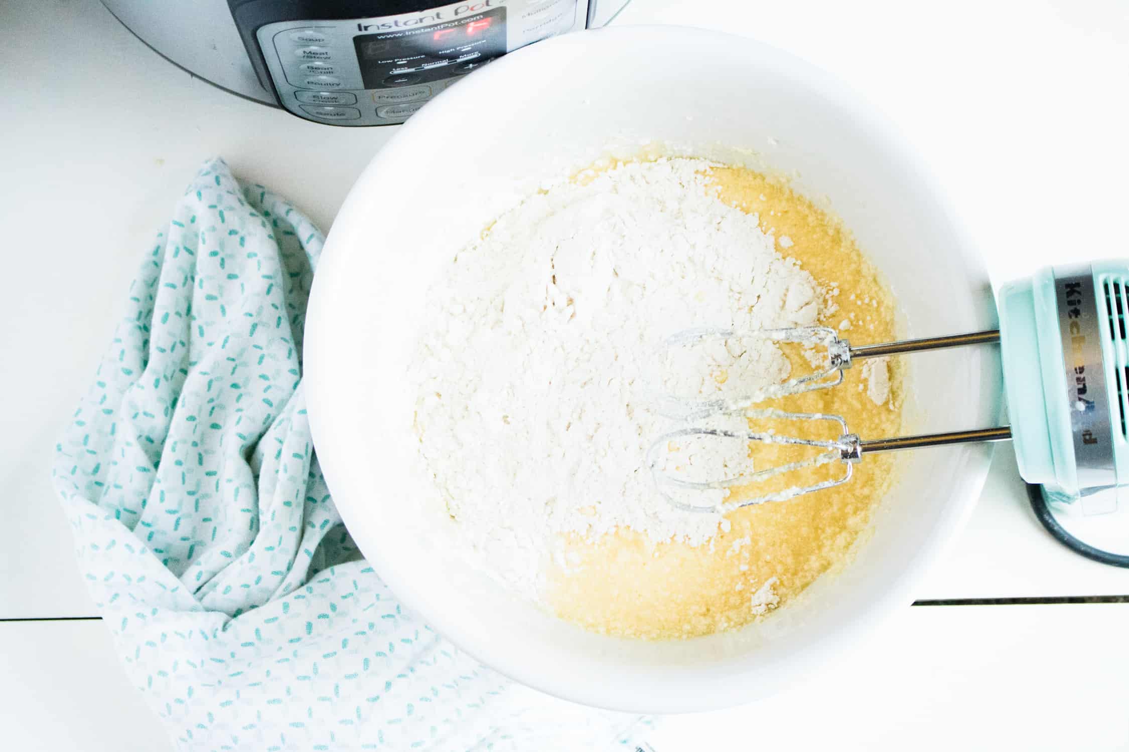 white ceramic bowl in foreground with ingredients for instant pot pound cake recipe being mixed by a turquoise hand mixer . There is a silver instant pot in back.