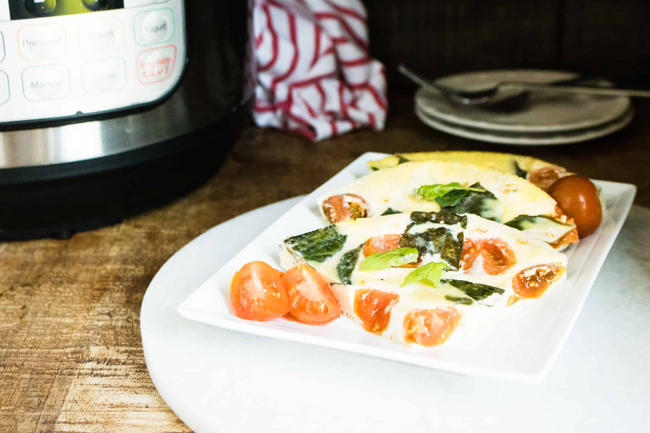 The finished Instant Pot Caprese Frittata served on a white platter and garnished with basil and cherry tomatoes next to the Instant Pot, a red striped napkin, two white lunch plates and two silver forks 