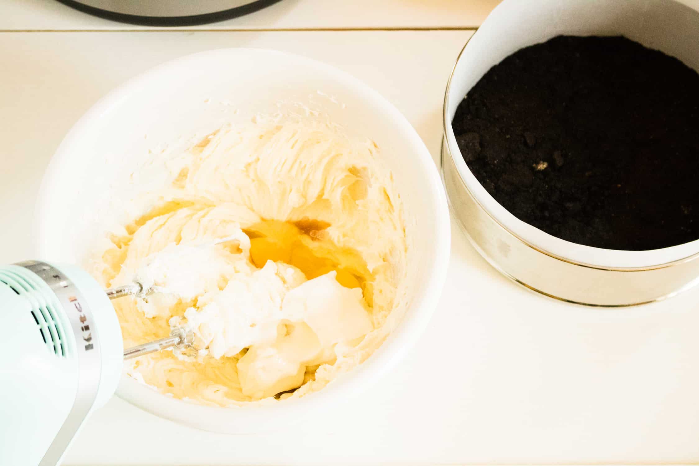 cream cheese, sugar and eggs in a white bowl being mixed together with a mixer next to a springform pan with an Oreo cookie crust