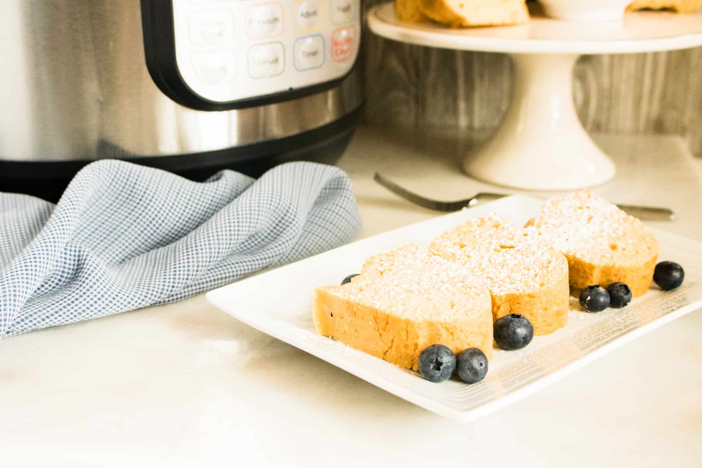 instant pot pound cake recipe on rectangular white plate in forefront with silver instant pot in background and white pedestal cake holder with cake on it to the right background