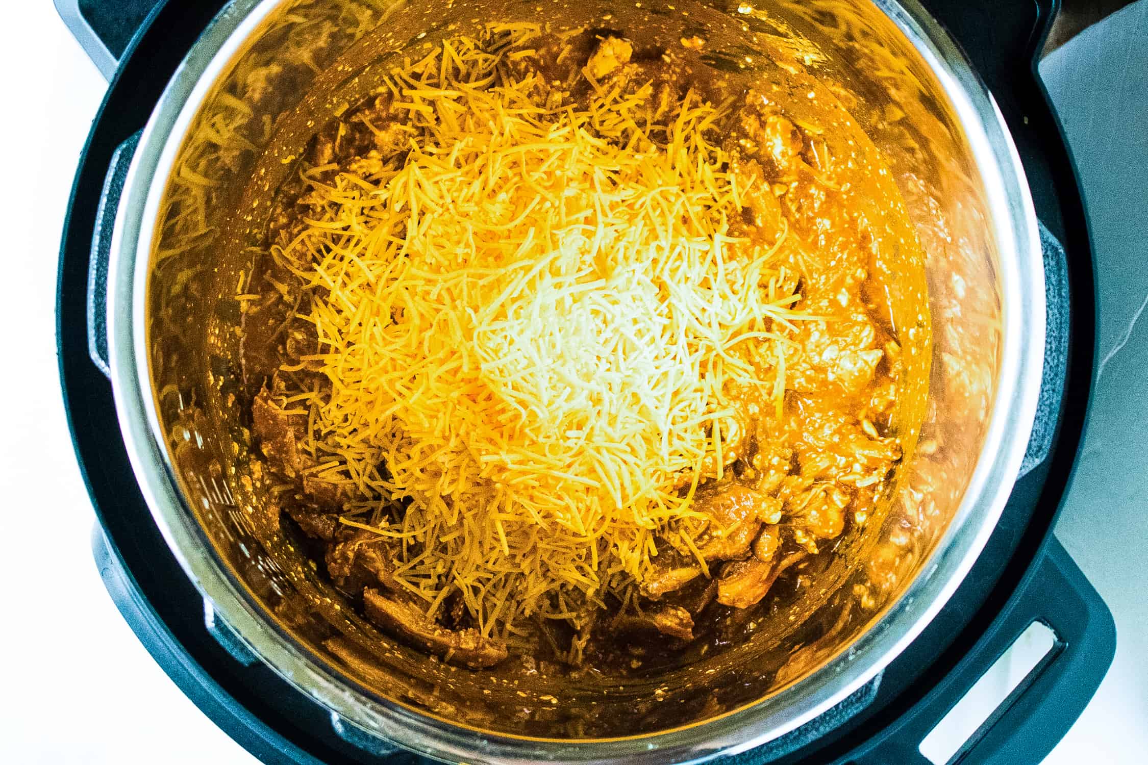 looking down into instant pot at ingredients for instant pot buffalo chicken dip with shredded cheddar cheese on top that has yet to be melted