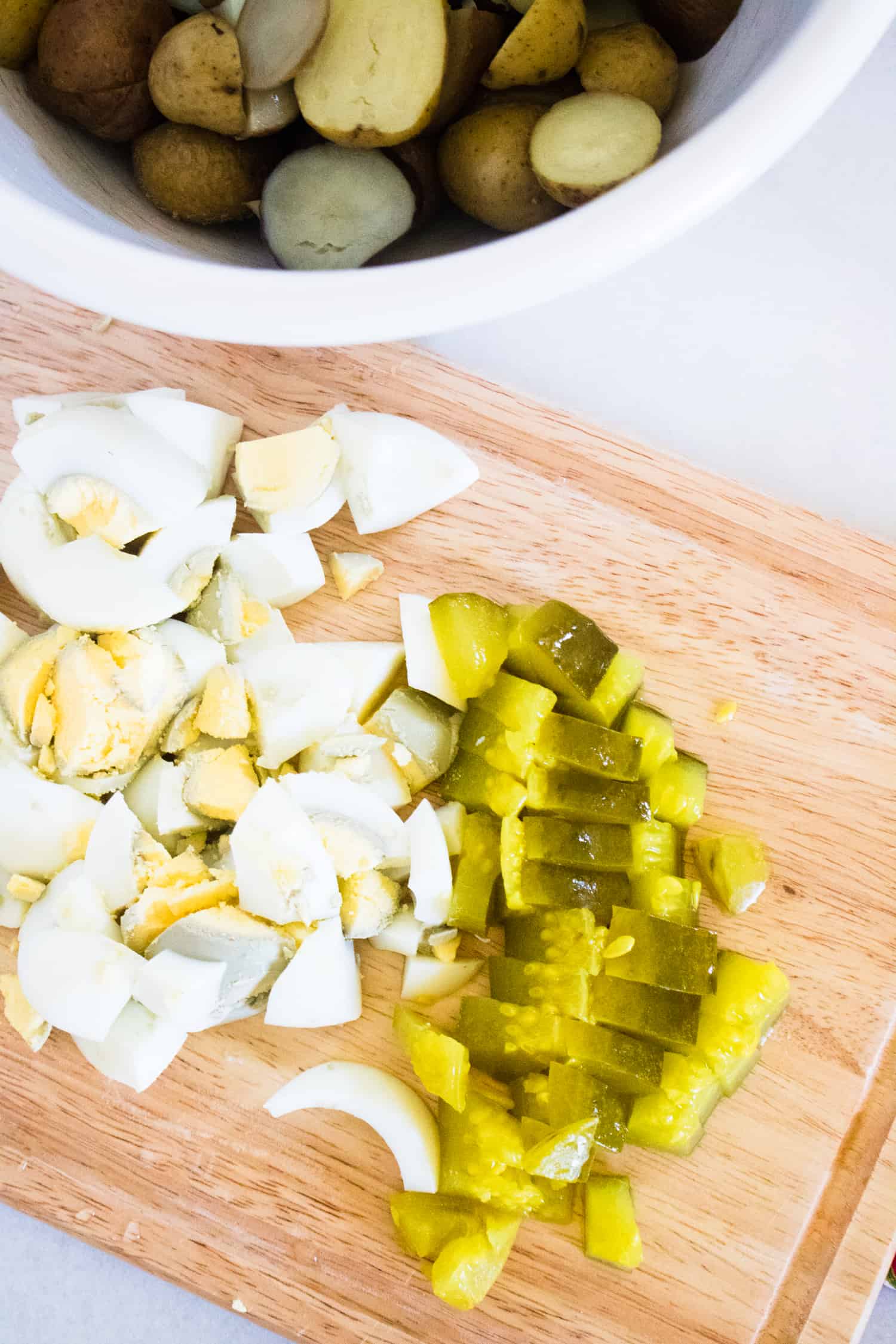 sliced potatoes in white bowl for instant pot heirloom potato salad in background with chopped hard boiled eggs and pickles on wooden cutting board in foreground