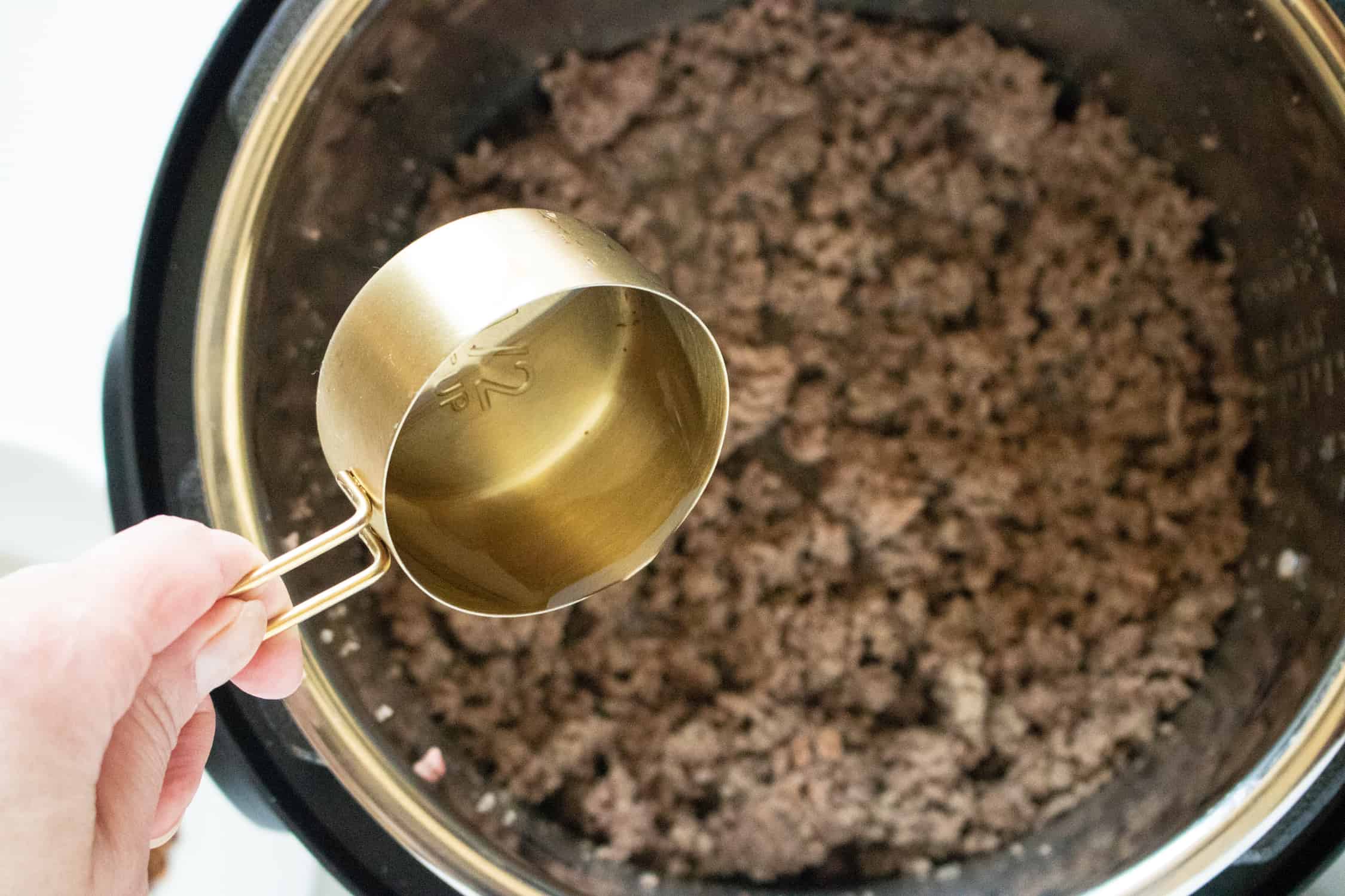 A hand pouring a cup of water into an Instant Pot filled with Instant Pot Taco Meat