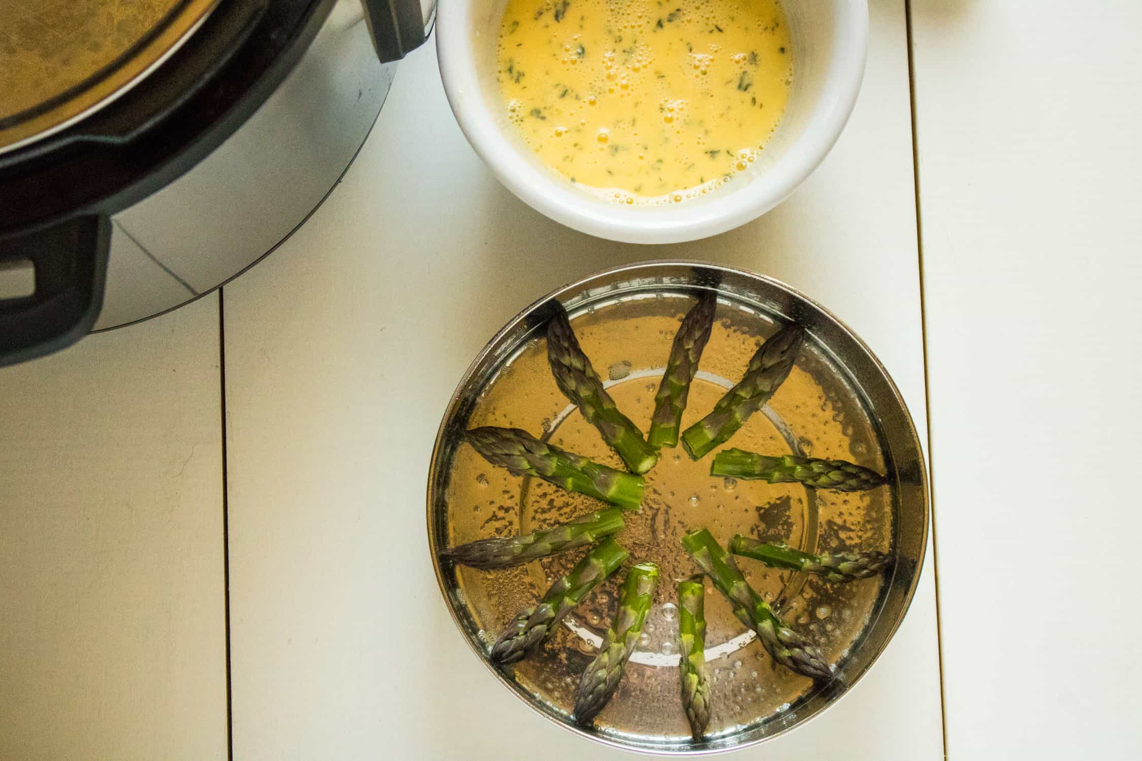 top-down view of asparagus tips arranged in the bottom of the 8-inch metal baking pan set next to the Instant Pot and the white bowl containing the Instant Pot Asparagus Parmesan Frittata raw egg mixture