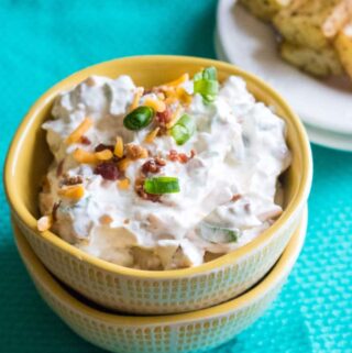 baked potato dip in small yellow bowl stacked on top of another yellow bowl on a table with a blue cloth