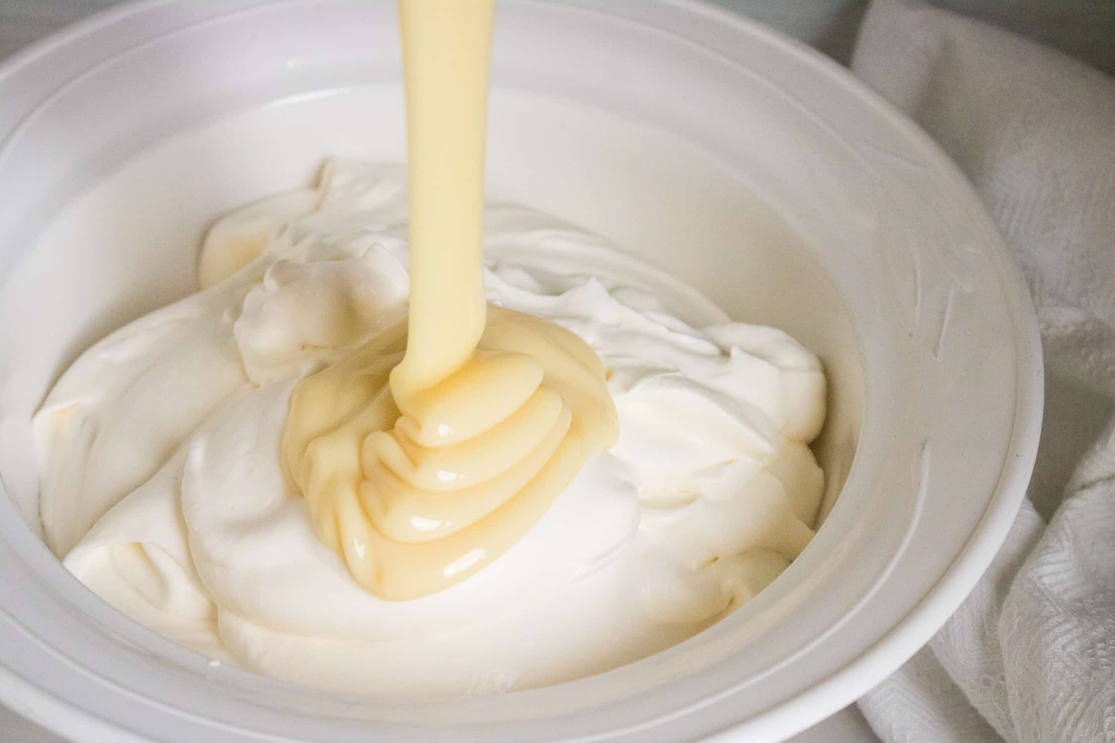 sweetened condensed milk being poured into a white mixing bowl filled with whipped heavy cream