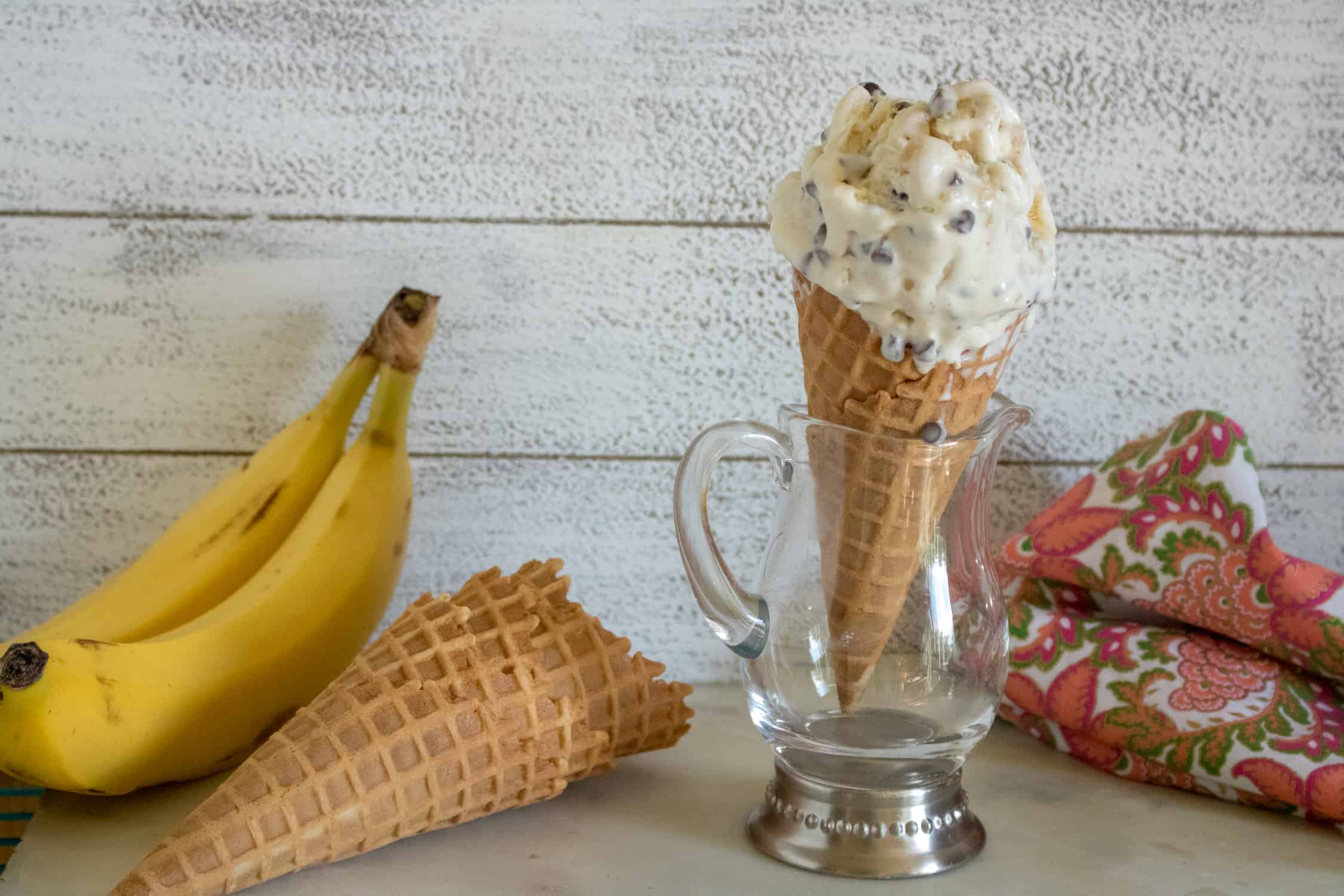 A waffle cone with a scoop of No-Churn Banana Chocolate Chip Ice cream standing in a glass cup on a table next to bananas, ice cream cones and a red patterned kitchen towel