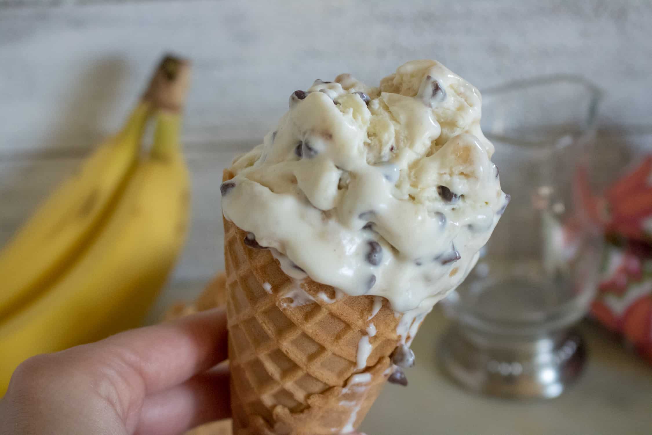 A waffle cone with a scoop of No-Churn Banana Chocolate Chip Ice cream being held up in the foreground, with two bananas, a glass cup and a red patterned kitchen towel in the background