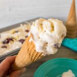 hand holding a scoop of black cherry white chocolate chunk no-churn ice cream on a sugar cone in front of a green dessert plate, cloth napkin and loaf pan containing the completed recipe