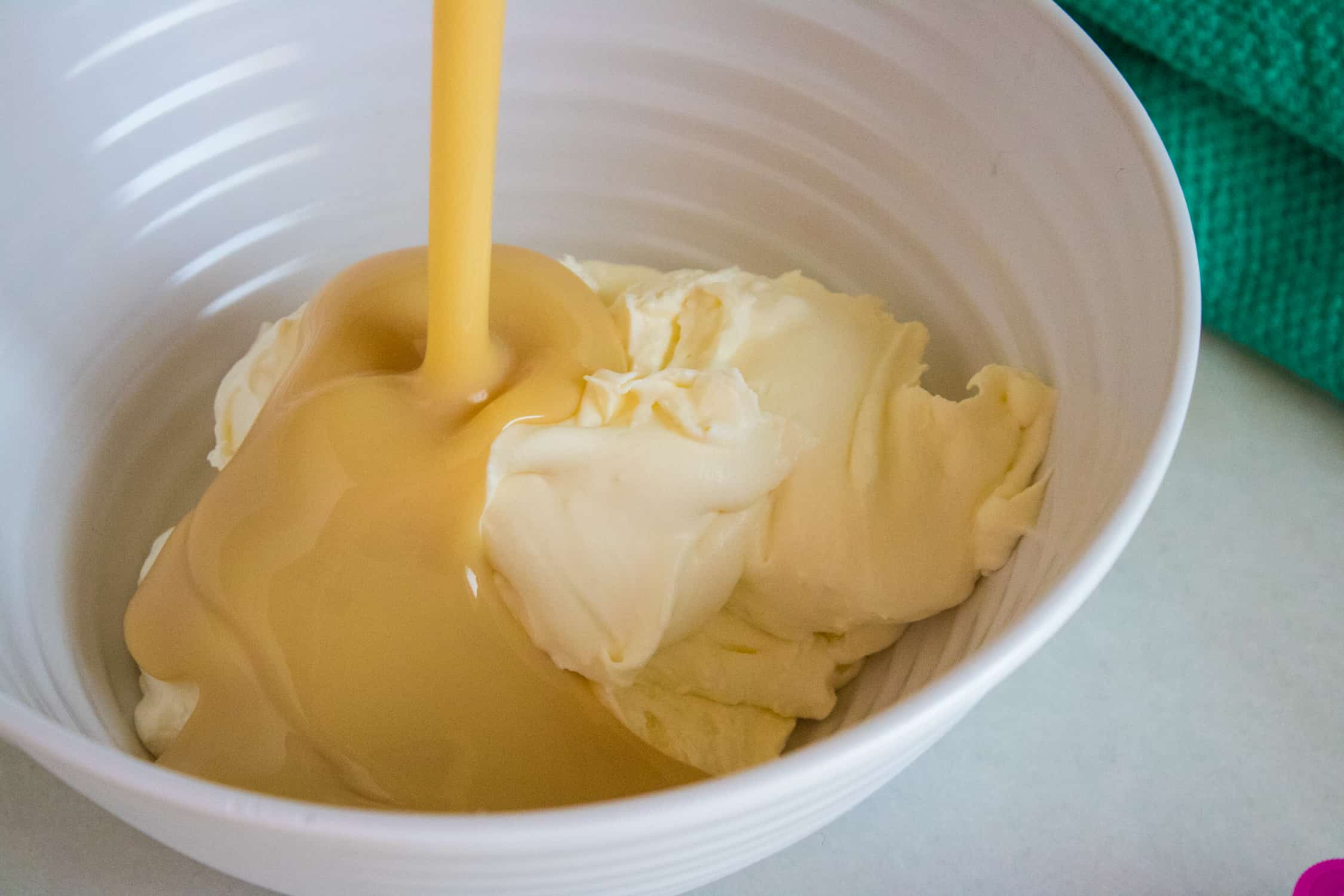 white ceramic bowl showing sweetened condensed milk being poured over whipped heavy whipping cream for bubble gum no-churn ice cream