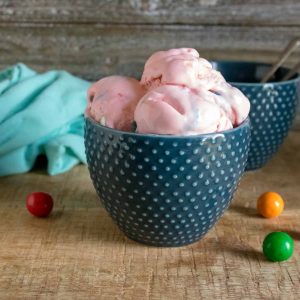 Bubble gum no-churn ice cream served in a blue polka-dot bowl next to a blue cloth napkin and multi-colored gum balls on a wood table