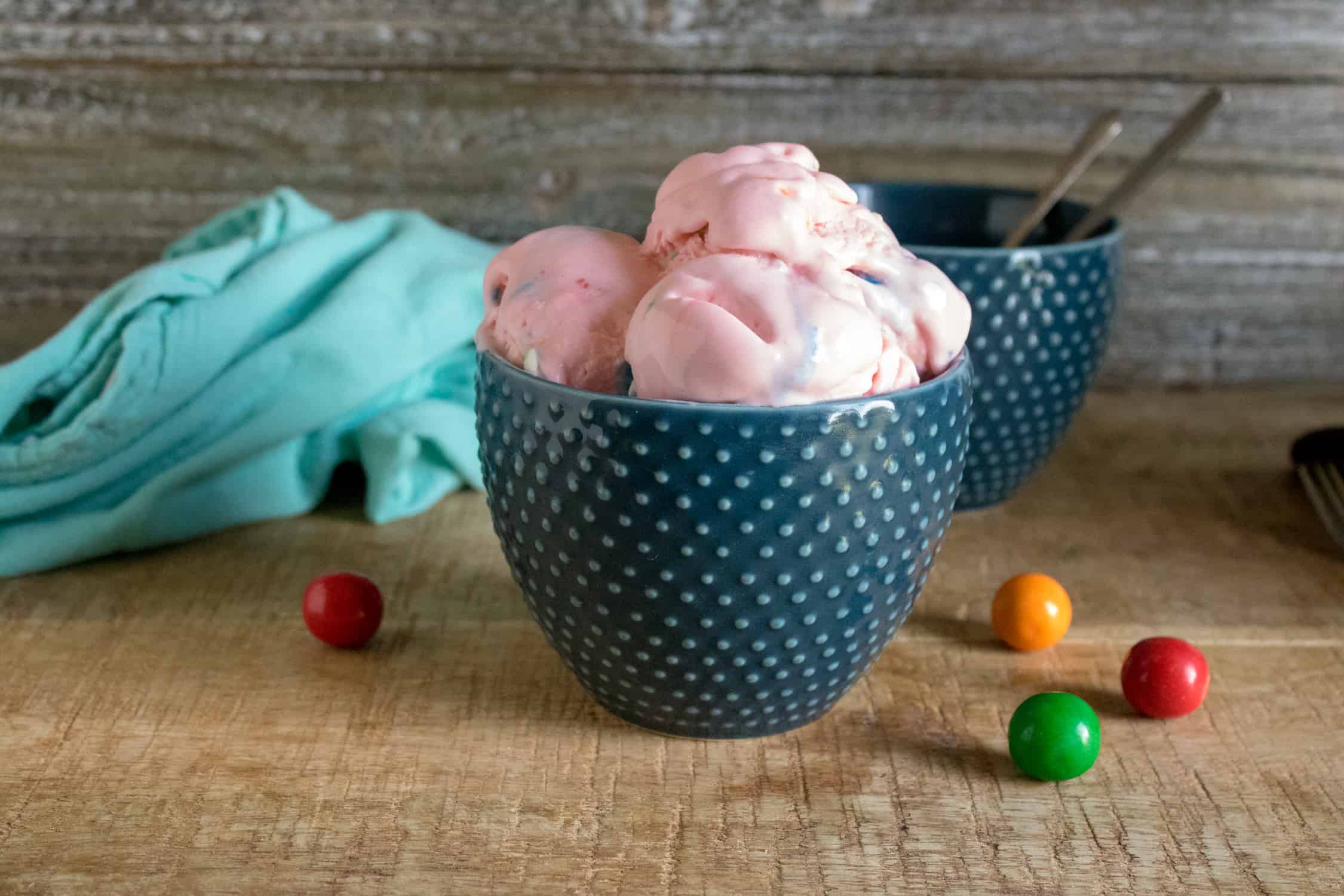 blue polka dot ceramic bowl with 3 scoops of pink bubble gum no-churn ice cream in it, blue towel in background and red orange and green gumballs scattered about