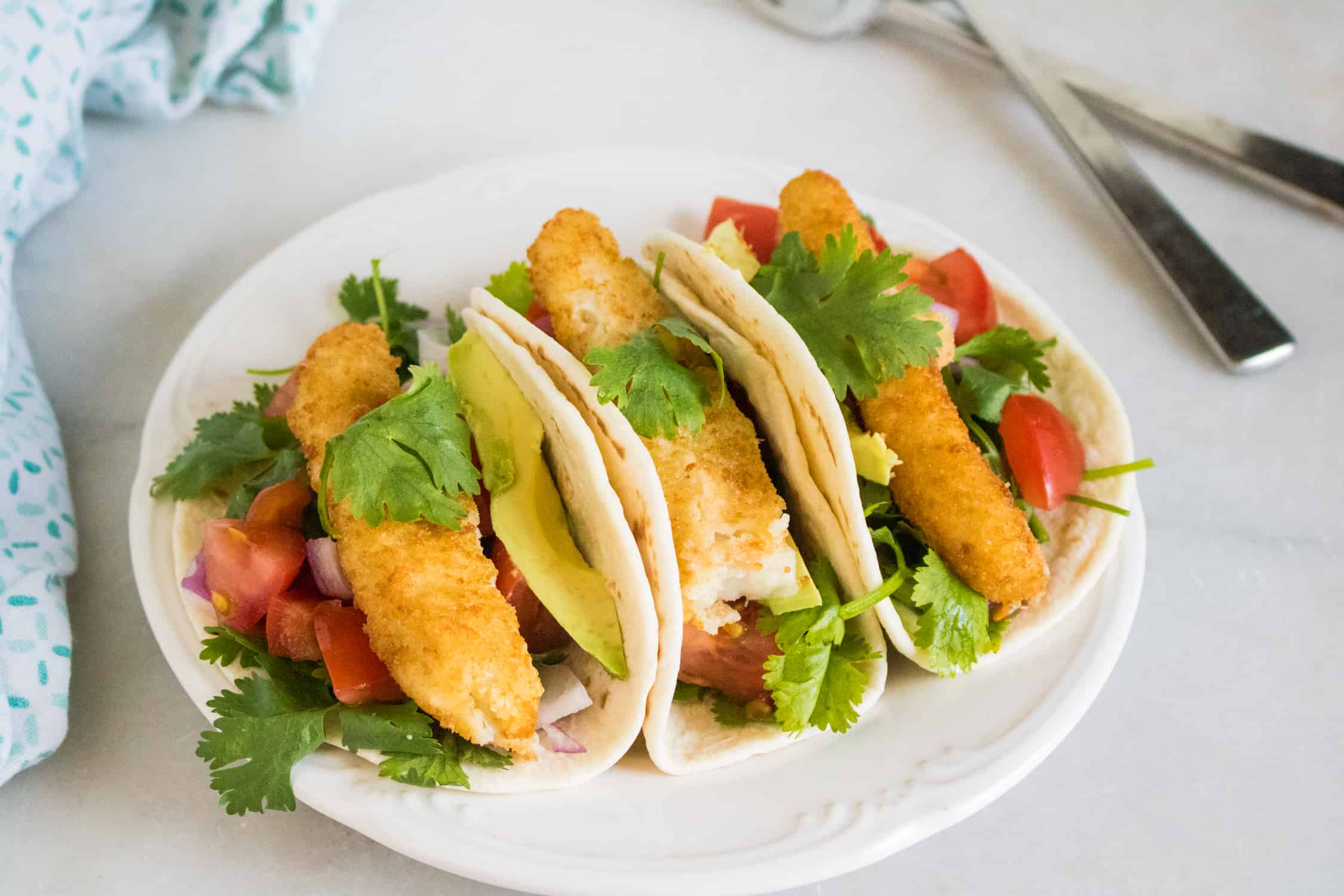top-down view of three crunchy fish tacos served on a white plate next to silver fork and blue print cloth napkin