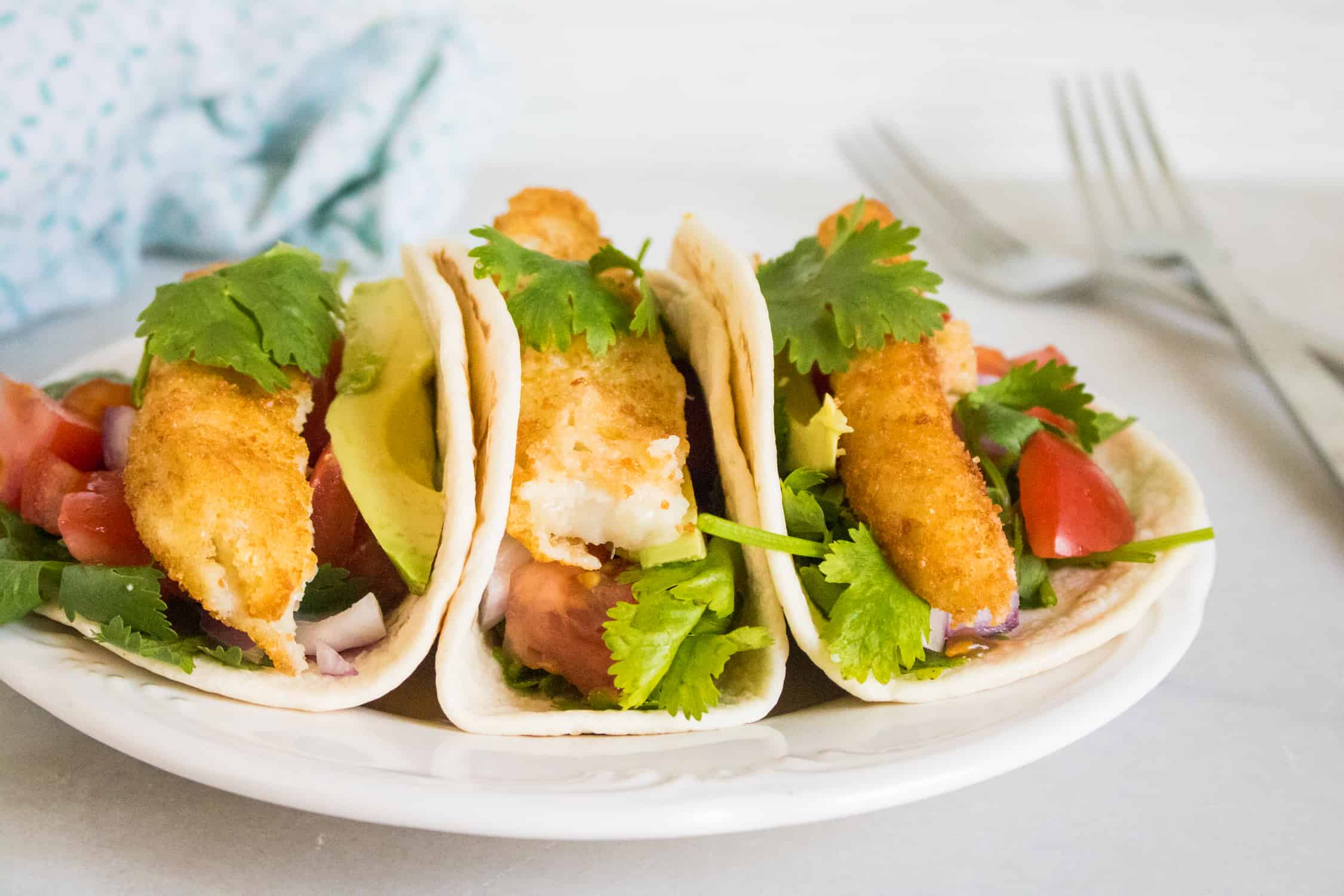 close up of the completed fish taco recipe featuring three tacos on a white plate set next to two silver forks and a blue print cloth napkin 