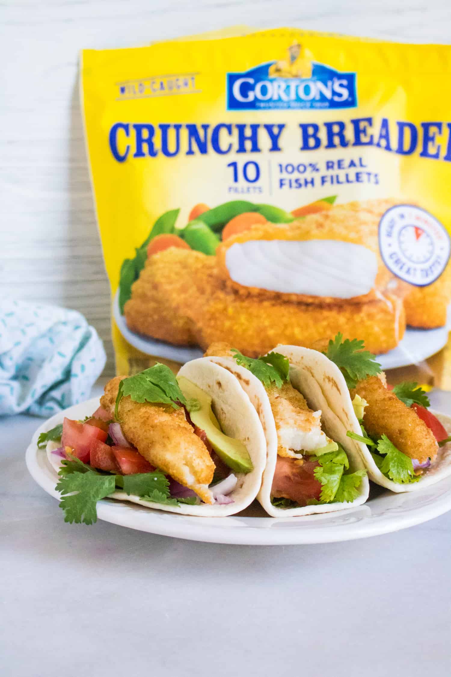 the completed fish taco recipe, featuring three crunchy fish tacos served on a white plate sitting in front of a bag of Gorton's Crunchy Breaded Fish Fillets
