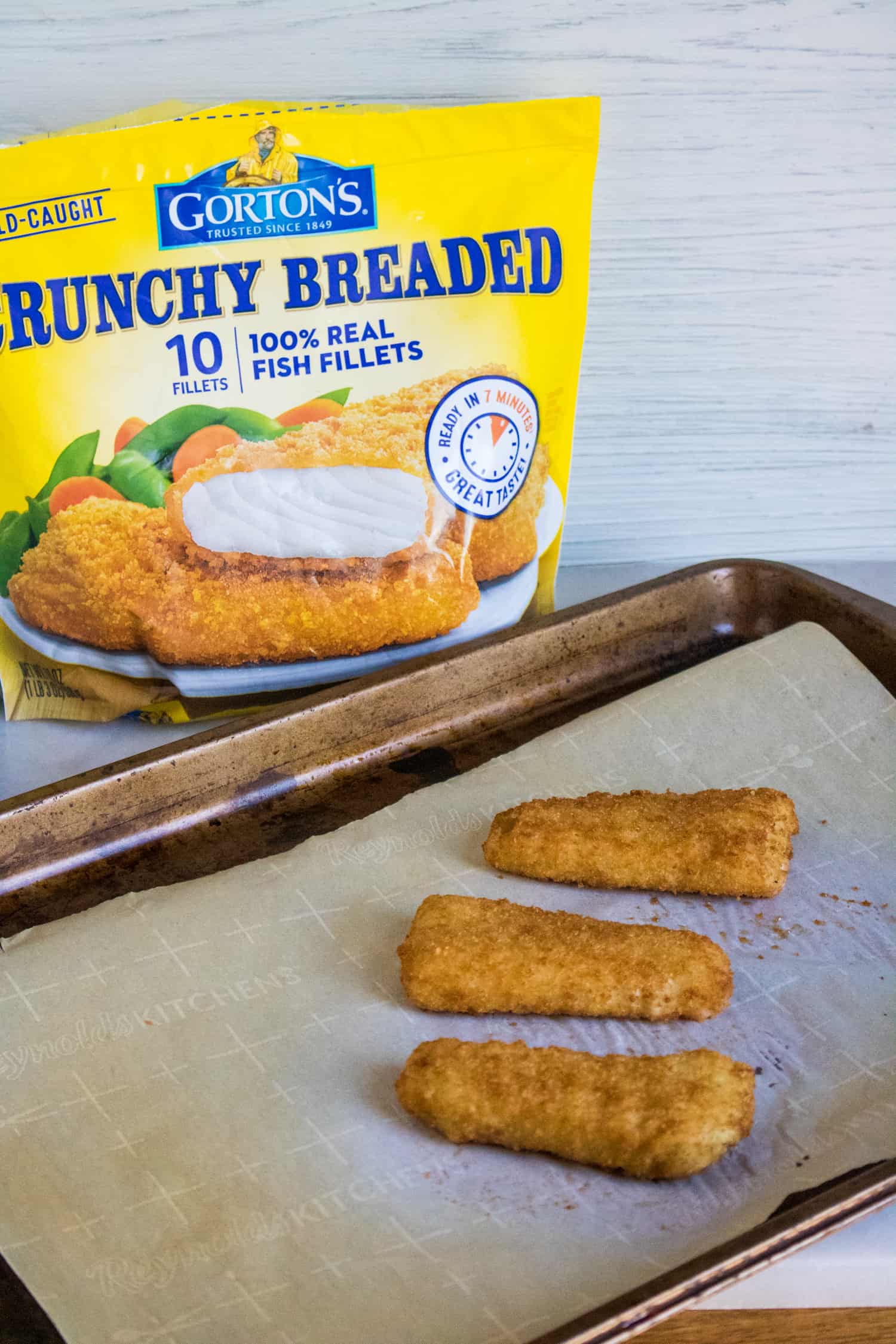 a bag of Gorton's Crunchy Breaded Fish Fillets sitting next to a baking sheet lined with parchment paper and three individual fish fillets set on top