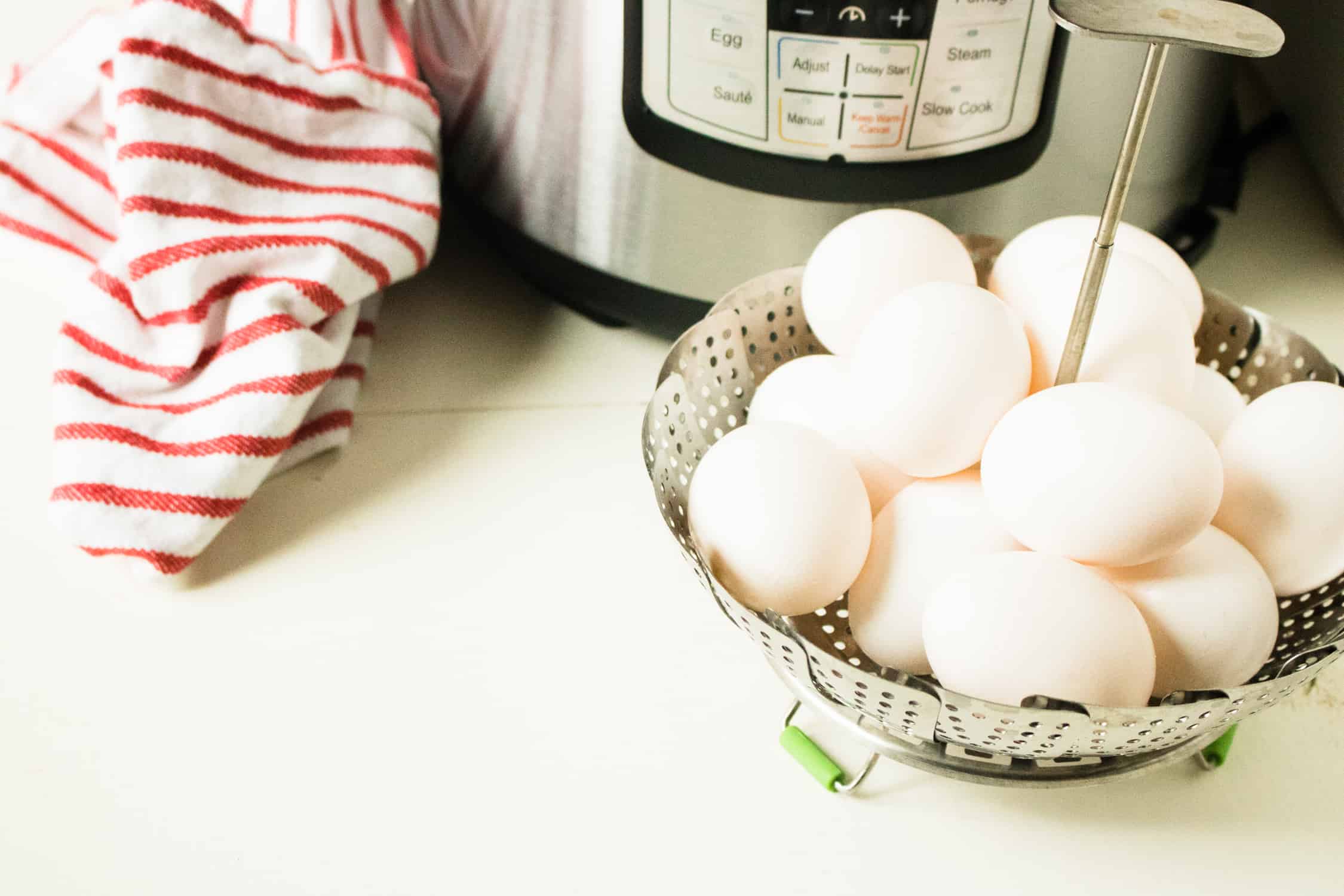 instant pot in background with 1 dozen eggs for Instant Pot Hard Boiled Eggs sitting in silver instant pot steaming basket with red and white disc cloth next to pot